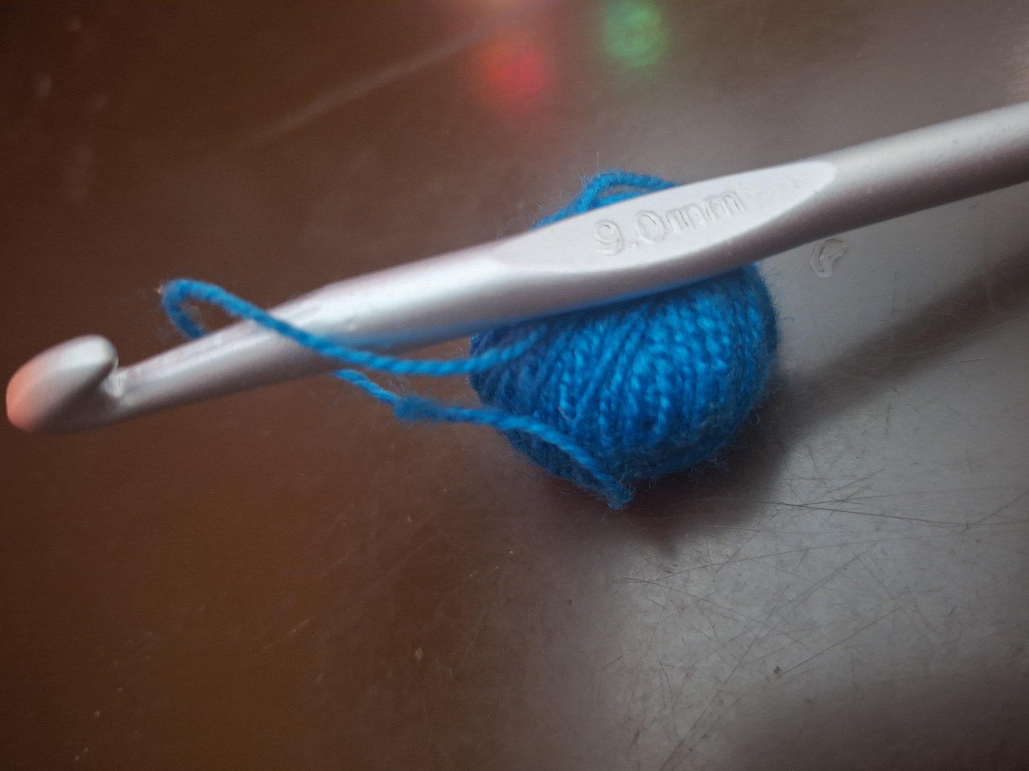 The image shows a close-up of a blue yarn ball and a silver crochet hook. The crochet hook is positioned diagonally across the yarn ball, with the yarn looped around it. The hook is marked with "9.0mm." The background is a blurry, reflective surface, possibly a table, with out-of-focus colorful lights visible in the upper portion of the image. There is no indication of the location being Kfar Saba, Israel. The scene is a still life of crafting materials.