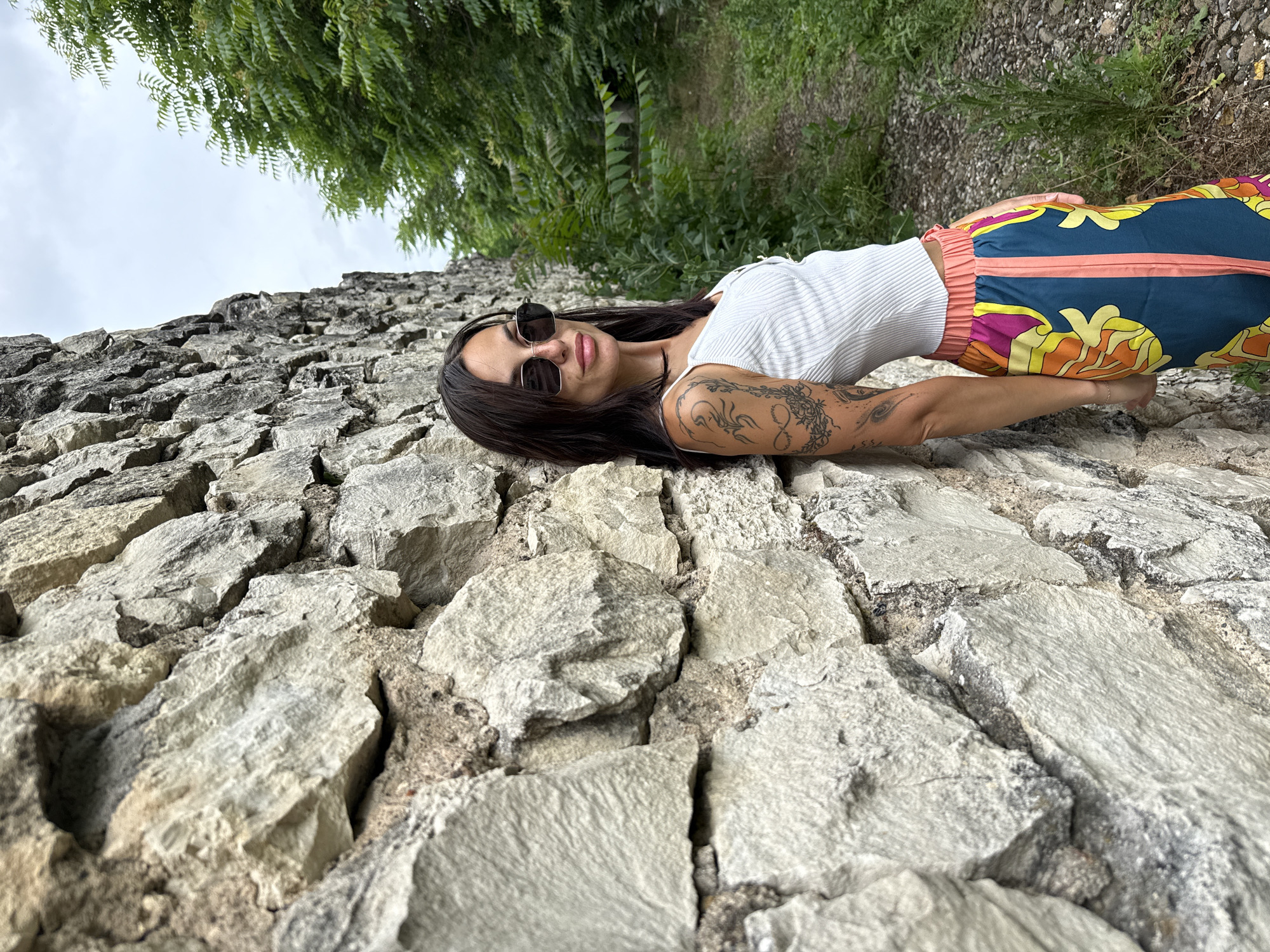 In Pleven, Bulgaria, a young woman with dark hair and sunglasses leans against a weathered stone wall. She is wearing a white tank top and colorful patterned pants. To her right, a gravel path is bordered by lush green foliage. The sky above is overcast.