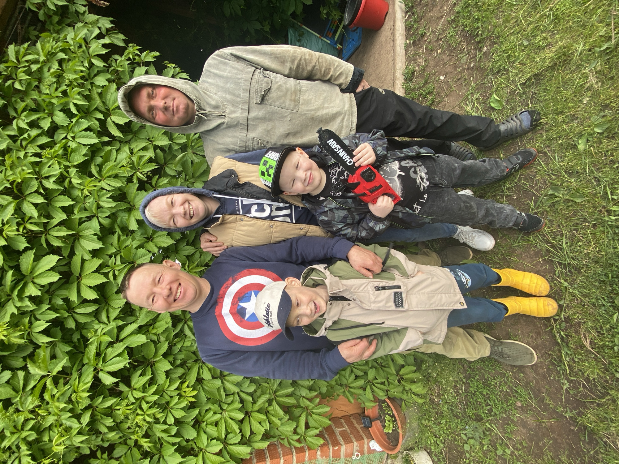 This outdoor photograph, taken in Chaikovsky, Russia, captures a multi-generational group of five males, likely a family, posing together. Two adult males are visible, one wearing a grey hooded jacket at the top, and another in a navy blue long-sleeved shirt featuring a prominent Captain America shield logo at the bottom. Between and around them are three children.
One child, positioned near the adult in grey, wears a blue hoodie layered under a tan jacket. Another child, centrally located, sports a camouflage jacket and a black baseball cap with green details, holding a red and black toy chainsaw labeled "CHAINSAW". The third child, nestled with the adult in the Captain America shirt, wears a light beige jacket with the text "JOHNHERO" and a light-colored baseball cap with "ATHLETIC COLLEGE TEAM" printed on it. The text "JSIC" is also visible on one child's shirt underneath a jacket.
All individuals are smiling broadly, conveying a sense of happiness and contentment. They are casually dressed in jackets, hoodies, and various footwear including black sandals, sneakers, and bright yellow rain boots.
The setting appears to be a garden or backyard, characterized by a dense backdrop of lush green, five-leaflet vines. The foreground features a mix of green grass and bare earth or a dirt path. A brick ledge with a small plant pot is visible towards the bottom right. In the background, a red bucket and a blue object can be seen. The lighting suggests it is daytime, possibly overcast, contributing to a soft and pleasant atmosphere.