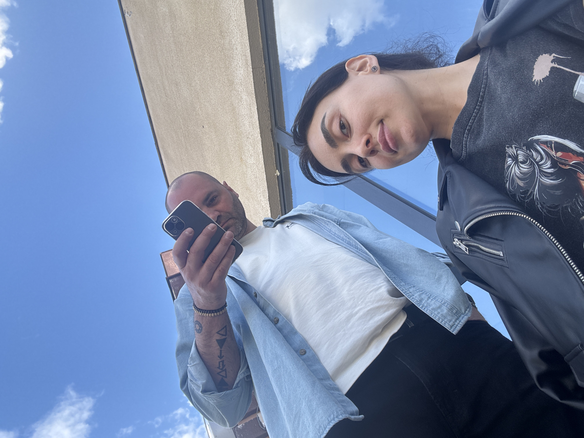 Two individuals are taking a selfie from a low angle, with the sky visible above. A man with a shaved head and a light beard is holding a smartphone, looking at the screen. He is wearing a white t-shirt, an unbuttoned light blue denim shirt, and black pants. He has a bracelet on his wrist and visible tattoos on his forearm. A woman with dark hair, pulled back from her face, is beside him. She is wearing a dark grey sweatshirt with a graphic design, and a black leather jacket. The background features a blue sky with some clouds and a beige awning. It is probable this photo was taken on the exterior patio of a location within Tbilisi, Georgia.