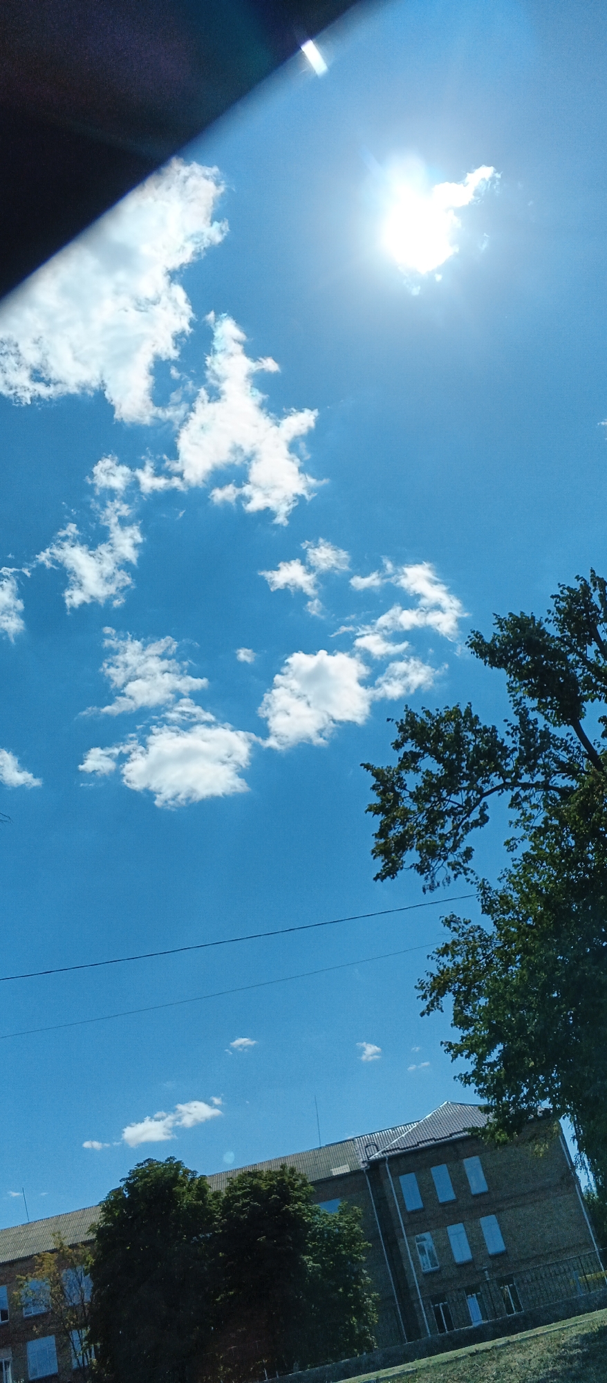 The image depicts a bright, sunny day with a clear blue sky dotted with fluffy white clouds. The sun, partially obscured by a cloud, is positioned in the upper right quadrant, casting a strong light. A dark, angular shape, possibly a vehicle roof or awning, enters the frame from the upper left, casting a shadow that partially covers a portion of the sky and some clouds.
In the lower portion of the image, a brick building with multiple windows is visible. Trees with green foliage are situated in front of and beside the building. A thin, dark power line stretches horizontally across the scene, above the building and below the trees. The perspective suggests the photo was taken from a slightly elevated position, looking towards the building and sky. The overall impression is an outdoor scene, likely in a built-up area given the presence of the building and power lines. The location is identified as Smila, Ukraine.