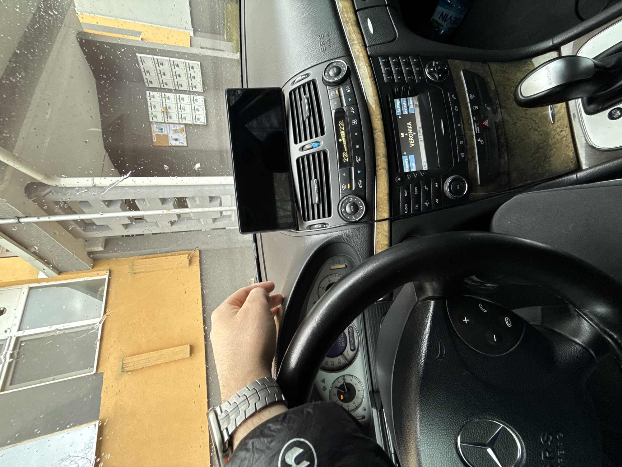 The image shows the interior of a Mercedes-Benz vehicle, with the driver's hand and watch visible. The car's infotainment system displays "VERONIKA" and "97.4 MHz," suggesting a radio station is playing. The climate control is set to 22 degrees Celsius. A tablet is mounted near the air vents. Through the rain-streaked windshield, one can see a building facade featuring windows and mailboxes. The speedometer shows a mileage of "887.9 km" and "P282978 S". Given the "VERONIKA" radio station and the location information provided, it's highly likely the scene is in Stara Zagora, Bulgaria.