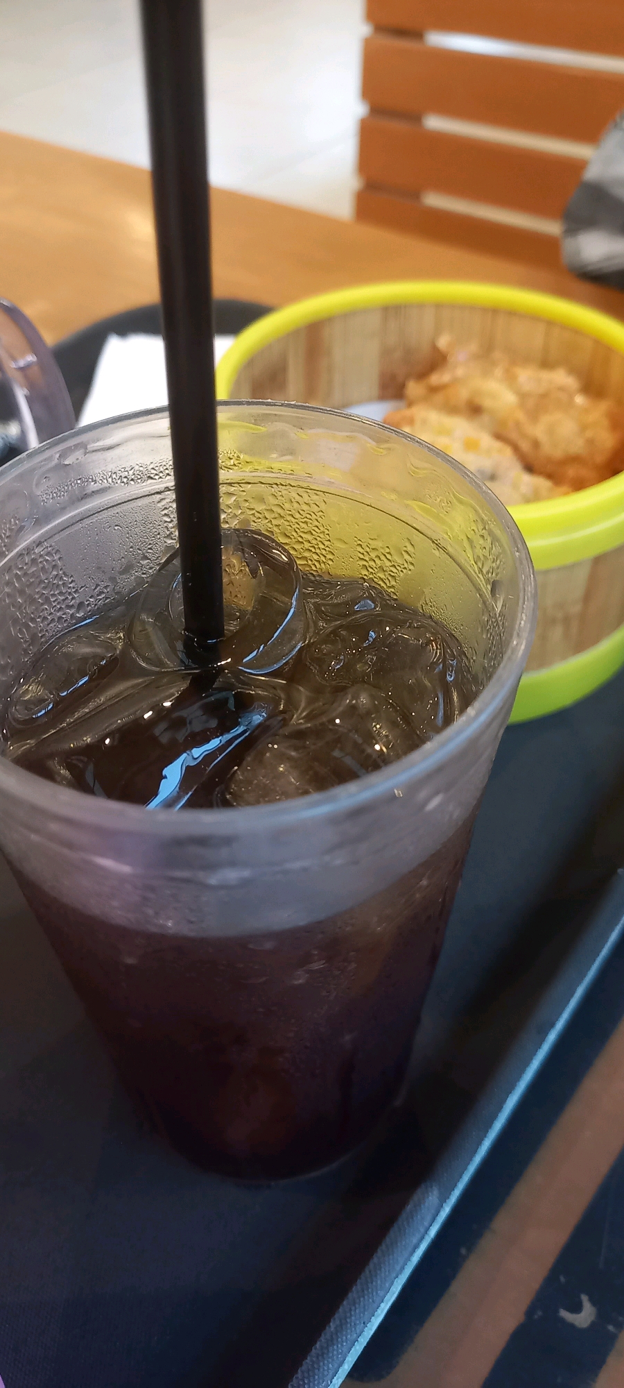 A close-up, eye-level shot focuses on a dark liquid in a clear plastic cup filled with ice and a black straw. Condensation covers the outside of the cup. In the blurred background, a yellow rimmed container holds food items. The scene appears to be set in an indoor dining environment.