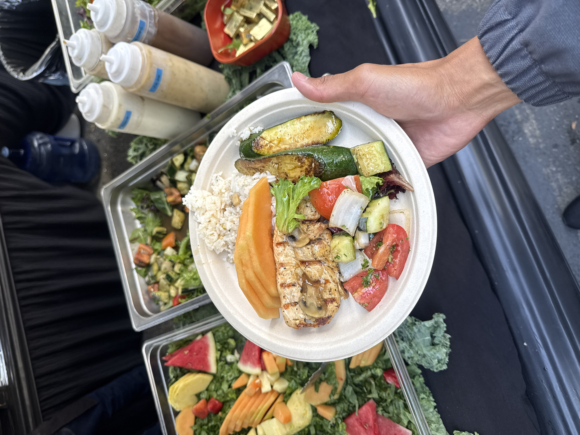 A top-down view captures a person's hand holding a plate of healthy food, set against the backdrop of an outdoor buffet or catering setup. The white, possibly compostable, plate is generously filled with a balanced meal: grilled chicken breast topped with sautéed mushrooms, a portion of rice (appearing to be brown or wild rice), several slices of orange cantaloupe, two large halves of grilled zucchini, and a fresh salad mix with diced tomatoes, cucumbers, and white onion pieces on a bed of greens.
In the background, a black tablecloth covers a surface, likely a table, on which various food items are displayed in metal serving trays and containers. To the upper left, four white squeeze bottles, presumably holding dressings or sauces, are visible in a metal holder. Blue labels are affixed to the bottles; one partially readable label says "GREEN G..." and another "JAPA...". To the upper right, a small red bowl contains golden, cubed items, possibly croutons or tofu. Below the main plate, two large metal serving trays are visible. One holds a salad with mixed vegetables, while the lower tray contains a fruit and greens salad featuring slices of watermelon and other fruit. Loose green kale leaves are scattered around the trays, suggesting a decorative or organic theme. A blue water bottle is visible on the ground in the lower-left corner, and parts of a person's arm and leg, clad in gray pants, are seen around the plate. The scene depicts a casual daytime outdoor dining event in Los Angeles, USA, offering a variety of fresh and prepared dishes.