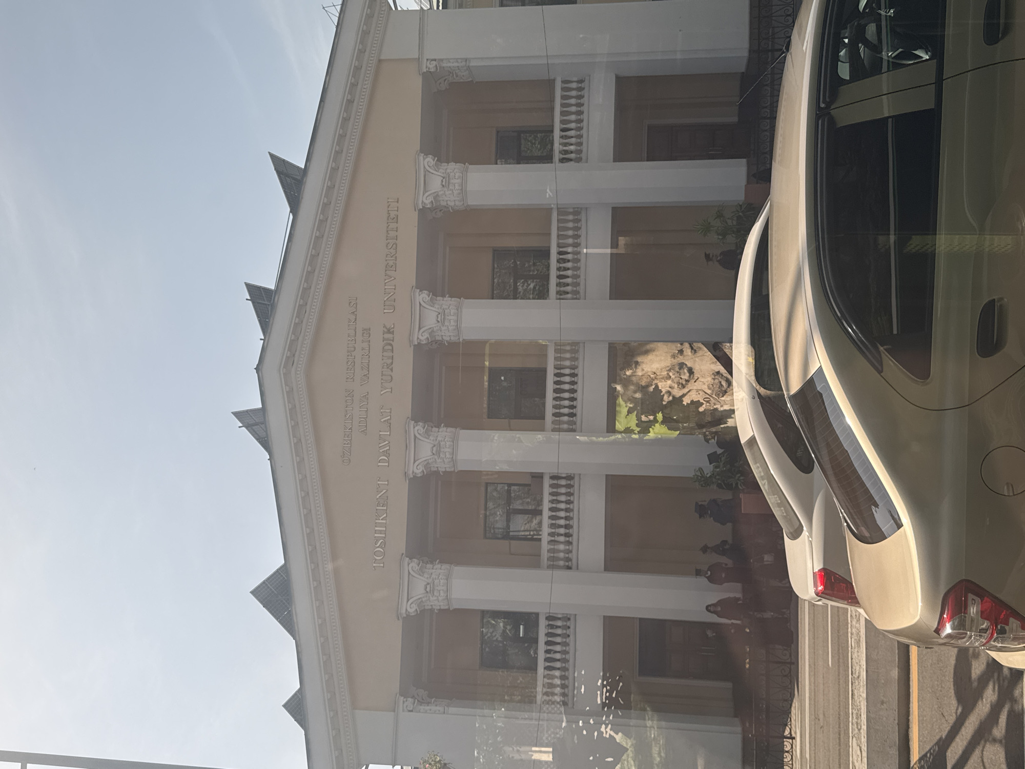 The image shows the front of a building with the inscription "O'ZBEKISTON RESPUBLIKASI ADLIYA VAZIRLIGI TOSHIKENT DAVLAT YURIDIK UNIVERSITETI" which translates to "REPUBLIC OF UZBEKISTAN MINISTRY OF JUSTICE TASHKENT STATE UNIVERSITY OF LAW".
The building has a classical architectural style with white columns and balustrades. The photo appears to have been taken from inside a car, as part of the vehicle is visible on the right side of the frame.
The location may be near the city of Kashgar mahalla, Uzbekistan, considering the sign's Uzbek language. However, the building itself is the Tashkent State University of Law in Tashkent, Uzbekistan.