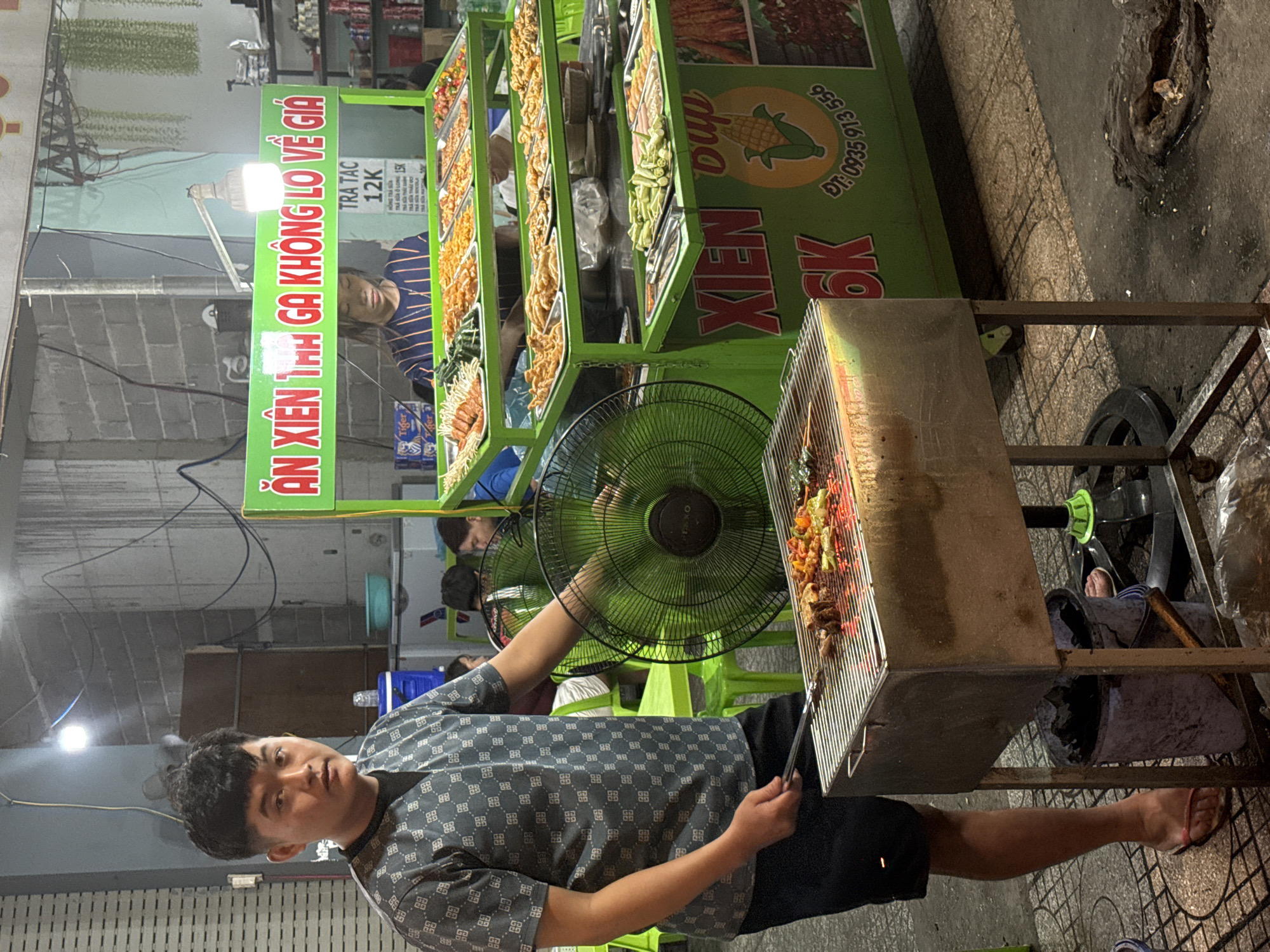 The image depicts an outdoor food stall, likely in Nha Trang Ward, Vietnam, specializing in grilled skewers. A man, wearing a dark patterned shirt and shorts, is seen tending to a charcoal grill laden with various grilled items, including meat and vegetables. To the right of the grill, a large green food cart is visible, displaying an assortment of pre-prepared skewers in metal trays. Signage on the cart, in Vietnamese, advertises "Xien Ga Khong Lo Ve Gia" which translates to "Chicken Skewers, Don't Worry About The Price." Another sign indicates prices, with "12K" and "15K" visible, likely referring to currency in thousands. A young woman, possibly an employee, is partially visible behind the food cart. A large, green industrial fan is positioned behind the man, likely to keep cool. The ground is paved with patterned tiles, and the overall setting suggests a street-side vendor.