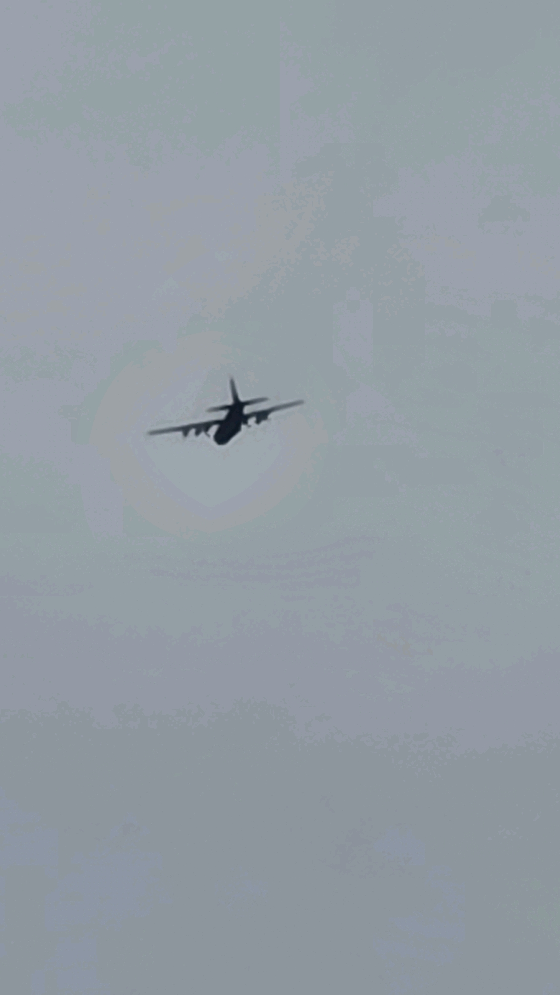 A silhouette of a C-130 Hercules aircraft is captured in flight against a hazy, light gray sky. The aircraft is oriented with its nose slightly upward and to the left, with its four engines and propeller blades visible. A faint circular halo effect, possibly from atmospheric conditions or lens flare, is discernible around the aircraft. The background is uniformly overcast and featureless. The image does not contain any information pertaining to the city of Dunnae-myeon, South Korea.
