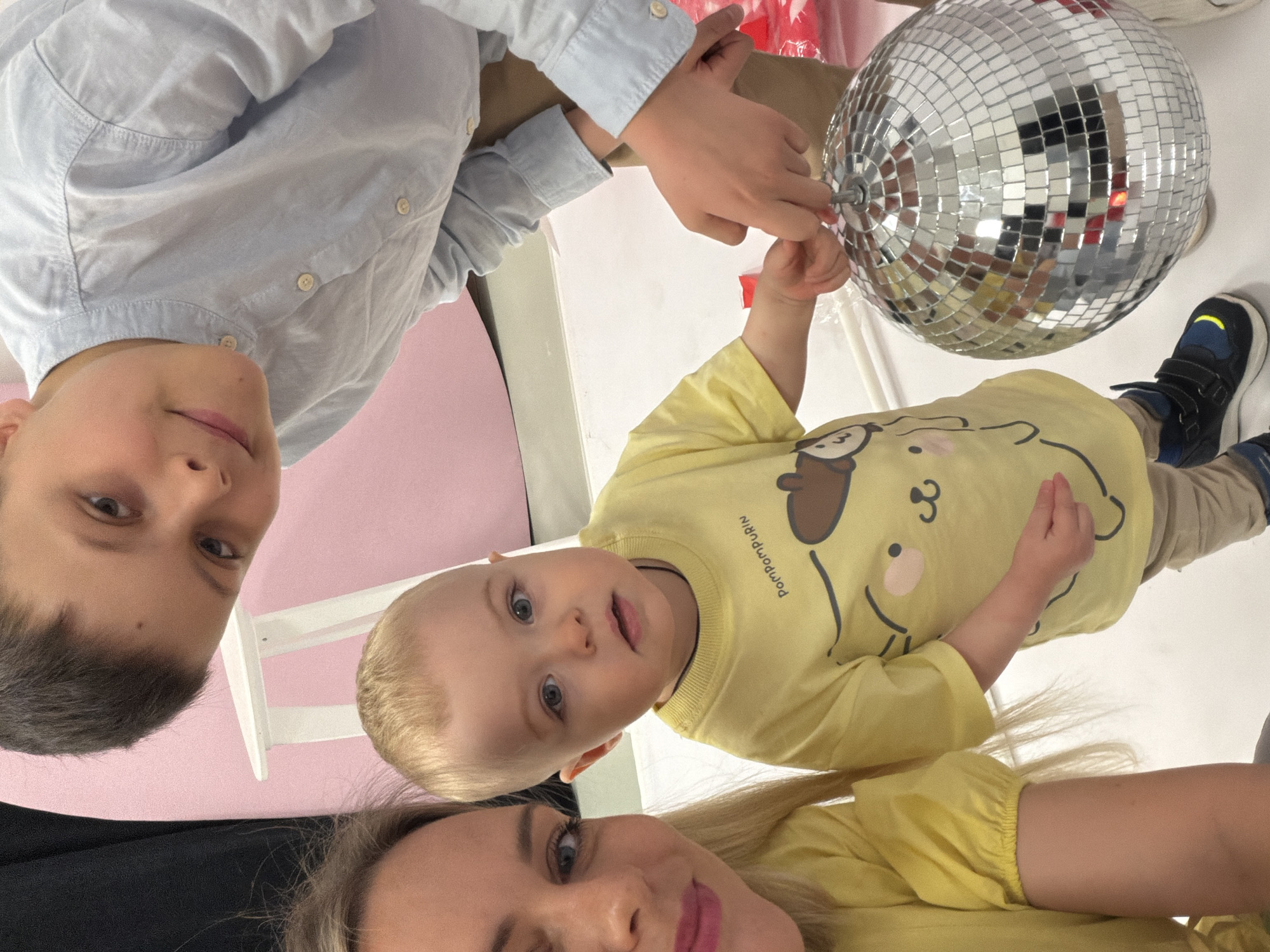 A group of three people, two children and one adult female, are holding a disco ball. The children are a young boy wearing a yellow shirt with a cartoon character on it, and an older boy wearing a light blue button-down shirt. The adult female has blonde hair and is wearing a yellow shirt. The background is a light pink color on one side and white on the other. A small white stool is partially visible. The location appears to be indoors. There is no specific information to confirm that the image was taken in Votkinsk, Russia.