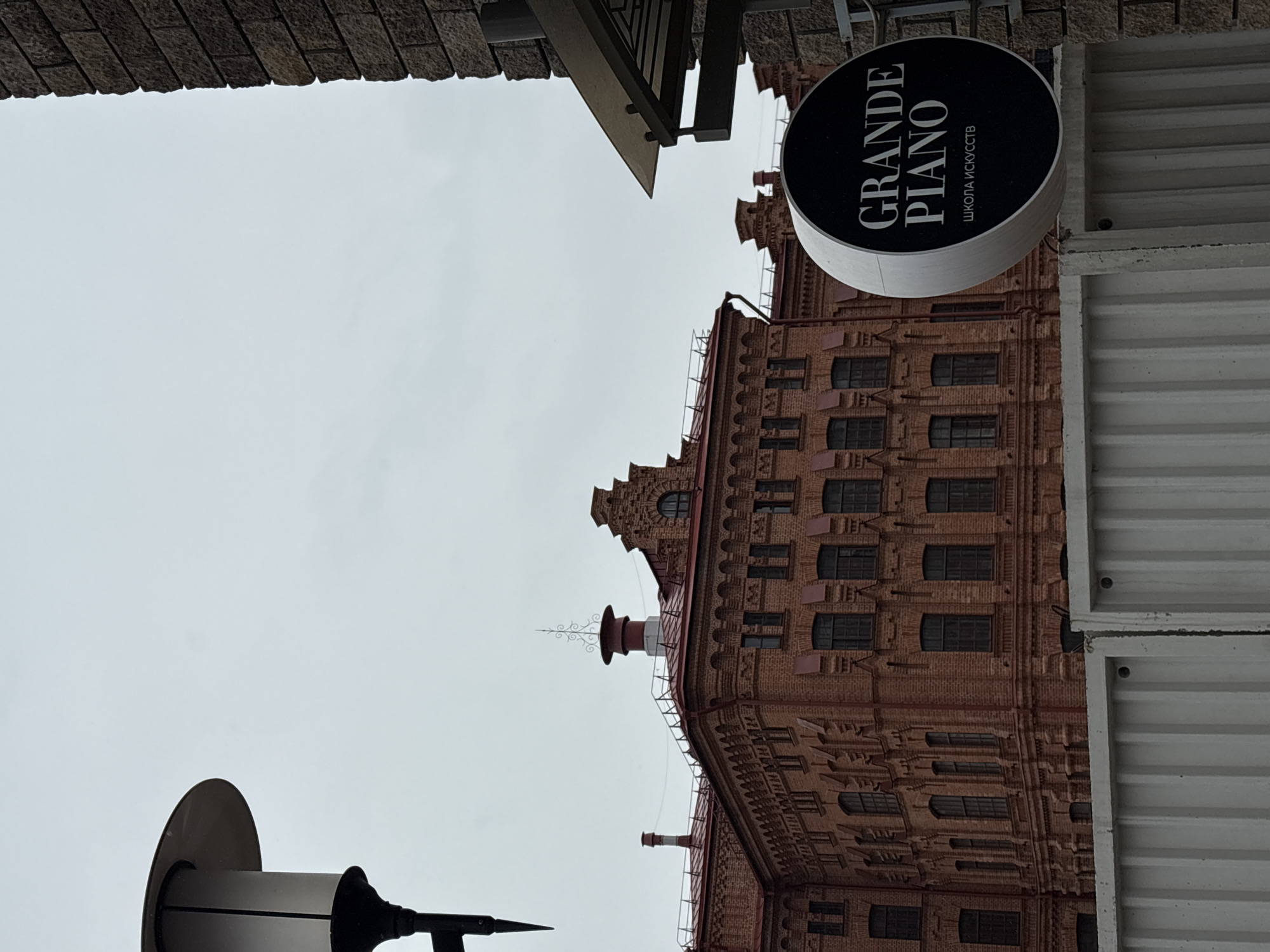 The image shows the exterior of a brick building with multiple windows, topped by architectural ornamentation and a red roof. A circular sign reading "GRANDE PIANO" in white letters is mounted on the building's right side. The sign includes the phrase "ШКОЛА ИСКУССТВ," likely indicating a school of arts. The sky is overcast. A brick wall is visible on the left side, and a corrugated metal structure is present on the right side. The scene is likely in Krasnoyarsk, Russia, based on the Cyrillic text on the sign. A street lamp is partially visible in the bottom left corner. The image is taken from a low angle, looking upwards.