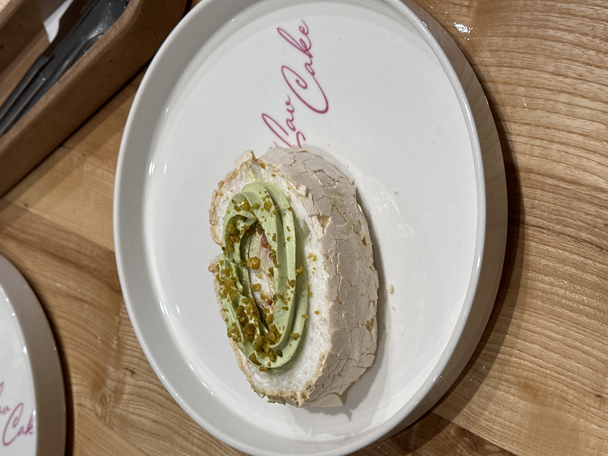 A close-up shot depicts a slice of meringue roll cake on a white plate, set on a wooden surface. The roll cake features a cracked meringue exterior and is filled with green cream and chopped pistachios. The plate is inscribed with the words "Pav Cake" in cursive pink font. To the left of the plate, a wooden utensil holder containing silverware is partially visible. Another similar plate is partially visible at the bottom left. The location is presumably a café or restaurant serving the "Pav Cake", although the specific city of Rostov-on-Don, Russia, cannot be verified from the image alone.