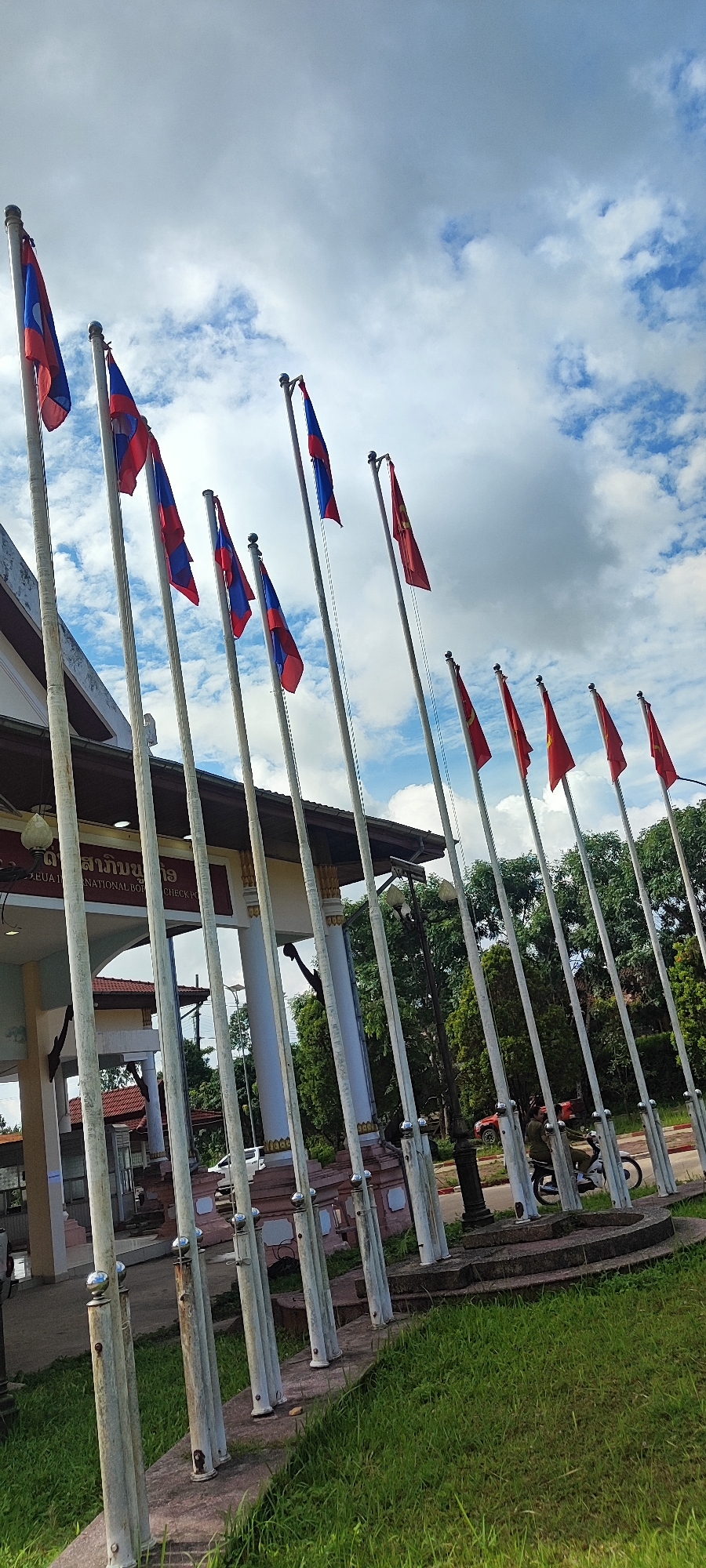 This image captures an exterior view of a border crossing in Xayden, Laos. A row of flagpoles, some with the Lao flag and others with the Vietnamese flag, stands prominently in the foreground, angled towards the sky. Behind the flagpoles, a building with traditional architecture, identified by signage in Lao script as a "NATIONAL BORDER CHECK POINT," is partially visible. The sky is partly cloudy, with patches of blue visible. Green grass covers the ground in front of the flagpoles, and a paved area with a road can be seen further back. A person on a motorbike is visible on the road. The overall scene suggests a functional border crossing with national symbols displayed.