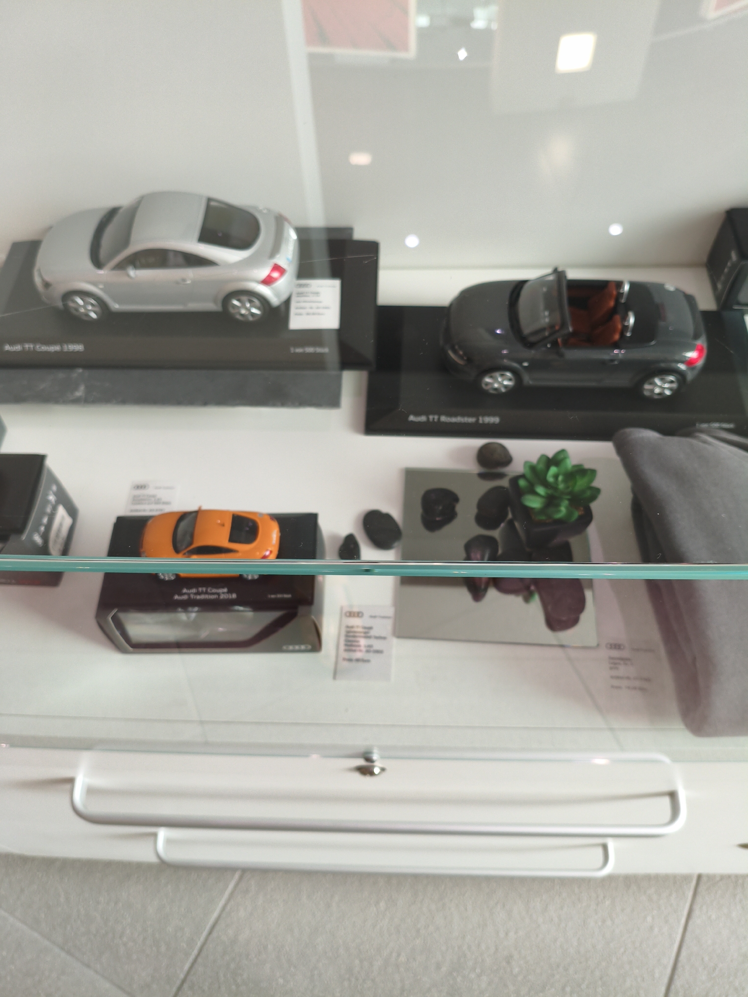 The image shows a display case containing several Audi model cars and some decorative items. In the top shelf, there is a silver Audi TT Coupe 1998 and a dark grey Audi TT Roadster 1999 with its top down and orange interior. Both models are on black display stands with labels. Below, on the second shelf, an orange Audi TT Coupe model sits on a black and silver display stand. To the right of the orange car, there is a small potted green succulent plant placed on a mirrored surface surrounded by several dark pebbles. A grey textile object is visible on the far right of the second shelf. The display case appears to be within a retail environment or a museum exhibit, possibly at the Audi Forum in Ingolstadt, Germany, given the brand's strong association with the city. The lighting in the scene is bright, suggesting an indoor setting.