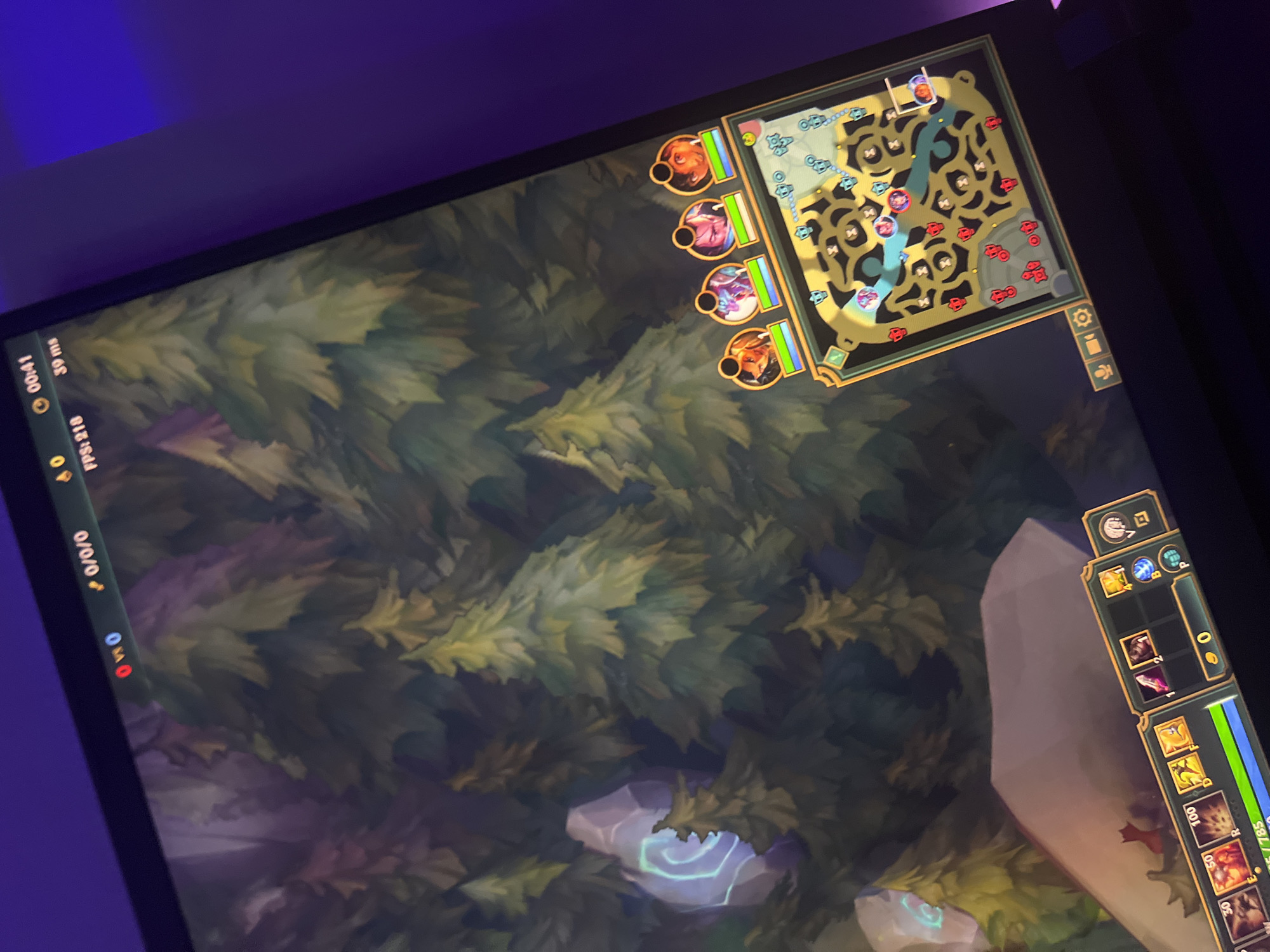 The image shows a close-up, angled view of a computer monitor displaying a game of League of Legends. The game is set in a forest environment. The top of the screen shows game information including the score (0/0/0), game time (00:41), FPS (218), and ping (39 ms). In the lower right corner, there is a mini-map of the game's terrain, showing player positions and objectives. Four character portraits are displayed above the mini-map, indicating the player's team. The bottom of the screen shows the player's inventory and abilities. The lighting in the image suggests the room is dimly lit with a purple hue, likely from ambient lighting. There is no indication of a city or specific location beyond the game environment.