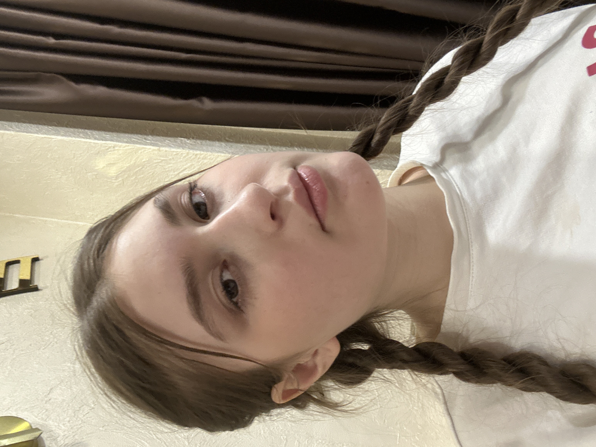 An indoor portrait taken in Sarapul, Russia, features a young female with light-medium brown hair styled into two visible braids. She faces the viewer directly, her gaze focused on the camera. Her skin appears fair with a slight flush on the cheeks, and her lips are slightly parted with a glossy appearance. She is wearing a plain white crew-neck t-shirt. The background consists of a light-colored, textured wall to her left and a portion of a brown, pleated curtain on her right. A gold-colored letter 'E' is partially visible on the upper left wall, acting as a decorative element. No other individuals or specific actions are depicted.