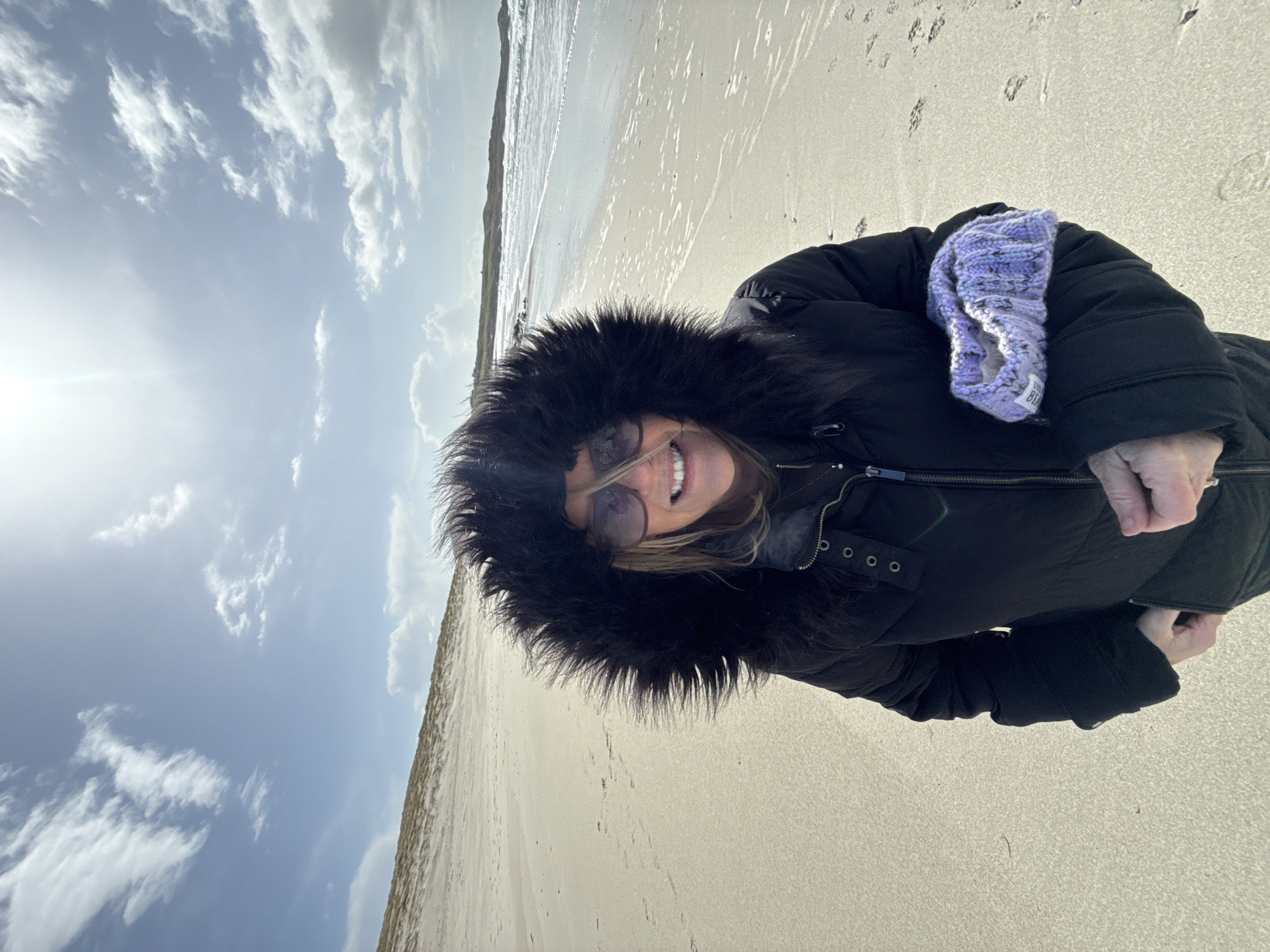 A woman in a black coat with a fur-lined hood is standing on a sandy beach. She is smiling at the camera and wearing sunglasses. The beach appears to be in an open area with a water body on the left side. The sky has a mix of clouds and sunshine. A purple item of clothing is tucked into her coat. There are footprints in the sand. The location is unknown.