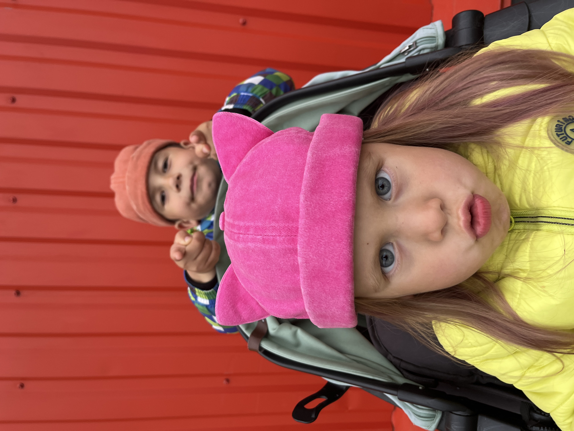 The media file shows two children in a stroller against a red corrugated metal wall. The child in the foreground, facing the camera, is wearing a bright yellow puffer jacket and a pink hat with cat ears. They have light-colored hair with streaks of pink and are looking at the camera with wide blue eyes and pursed lips. The child in the background, slightly out of focus, is wearing an orange beanie and a multi-colored plaid jacket. They are looking towards the camera and holding something in their hands.
The image appears to be taken outdoors during the daytime, with soft lighting that suggests an overcast sky. The red wall provides a vibrant backdrop and is the dominant element of the setting. The stroller is partially visible, with its light green lining and black frame.
The overall mood is casual and candid, capturing a moment of the children in the stroller. The expressions on their faces are curious and playful. There is no visible text in the image.