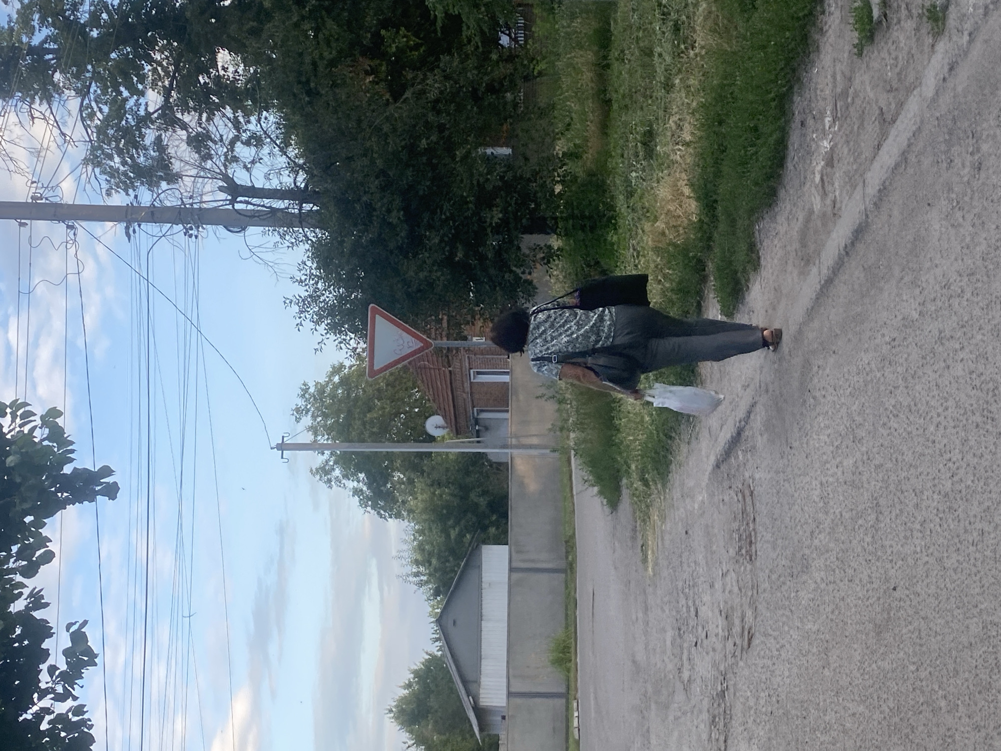 In Poltava, Ukraine, a woman walks away from the camera on a paved road with patches of gravel and dirt. She is carrying a white plastic bag in her right hand and a dark tote bag over her left shoulder. She is wearing a patterned short-sleeved shirt and dark pants. To her left is a low concrete wall with a building behind it, and further on, a larger building with a grey roof. To her right, there is a grassy verge and a traffic sign is visible on a pole. The sky is blue with scattered clouds. The scene appears to be in a rural or suburban area.