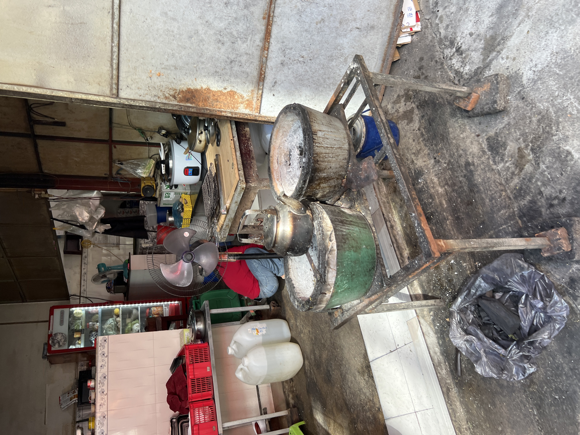 The image captures an outdoor food stall in Nha Trang, Vietnam, characterized by its utilitarian and somewhat disorganized setup. A person, clad in a red shirt and blue jeans, is crouched down in the background, attending to some unseen task. Dominating the foreground are two large cooking pots, likely used for charcoal grilling or boiling, positioned on a metal stand. One pot is green and the other is metallic, both appearing well-used with ash and remnants of cooking. Nearby, a plastic bag filled with what appears to be charcoal lies on the ground. The stall is equipped with various cooking implements and storage units, including red plastic crates, large water containers, a refrigerator, and a rice cooker. The overall environment suggests an active, albeit rustic, culinary workspace.