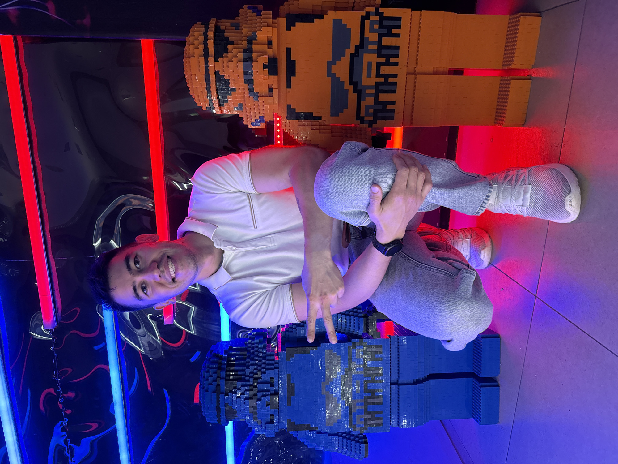 The image shows a man posing next to two LEGO figures. He is squatting between them and holding up a peace sign. The LEGO figures are orange and blue and stand upright. The man is wearing a white polo shirt, jeans, and white sneakers. He has a watch on his left wrist. The background is a wall with blue and red neon lights. The floor appears to be tiled. The image was taken indoors in Kazan, Russia.