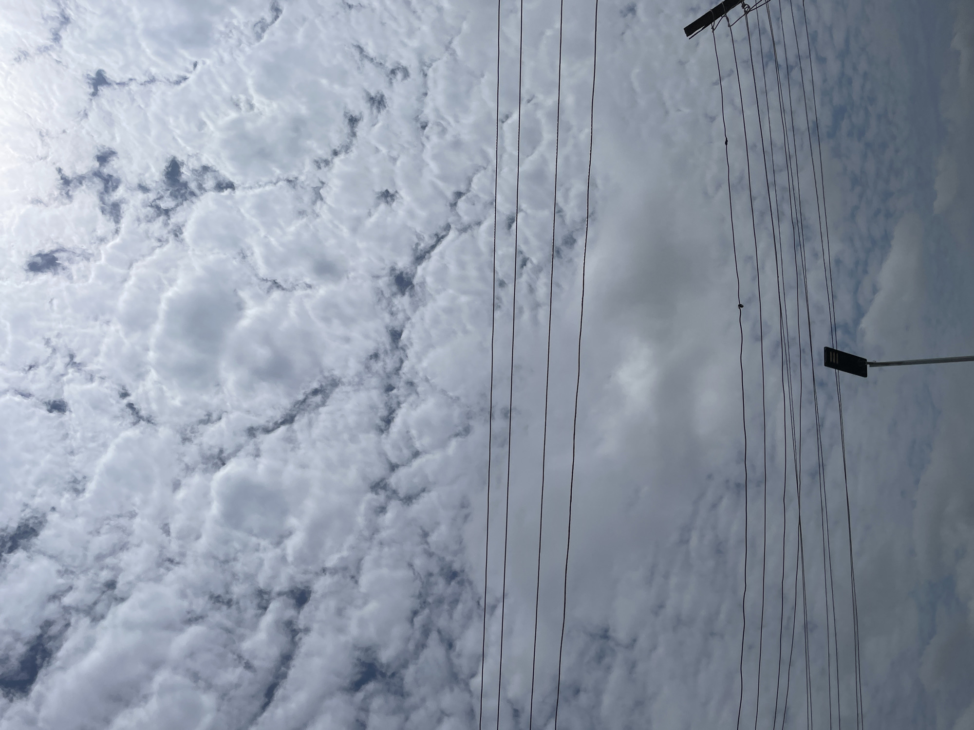 A streetlamp stands tall against a sky filled with fluffy, white clouds and patches of dark gray. Multiple power lines stretch horizontally across the frame, obscuring parts of the sky. A wooden pole with wires is visible on the right side of the image. The scene suggests an outdoor setting in Barakin Lerawi, Nigeria.