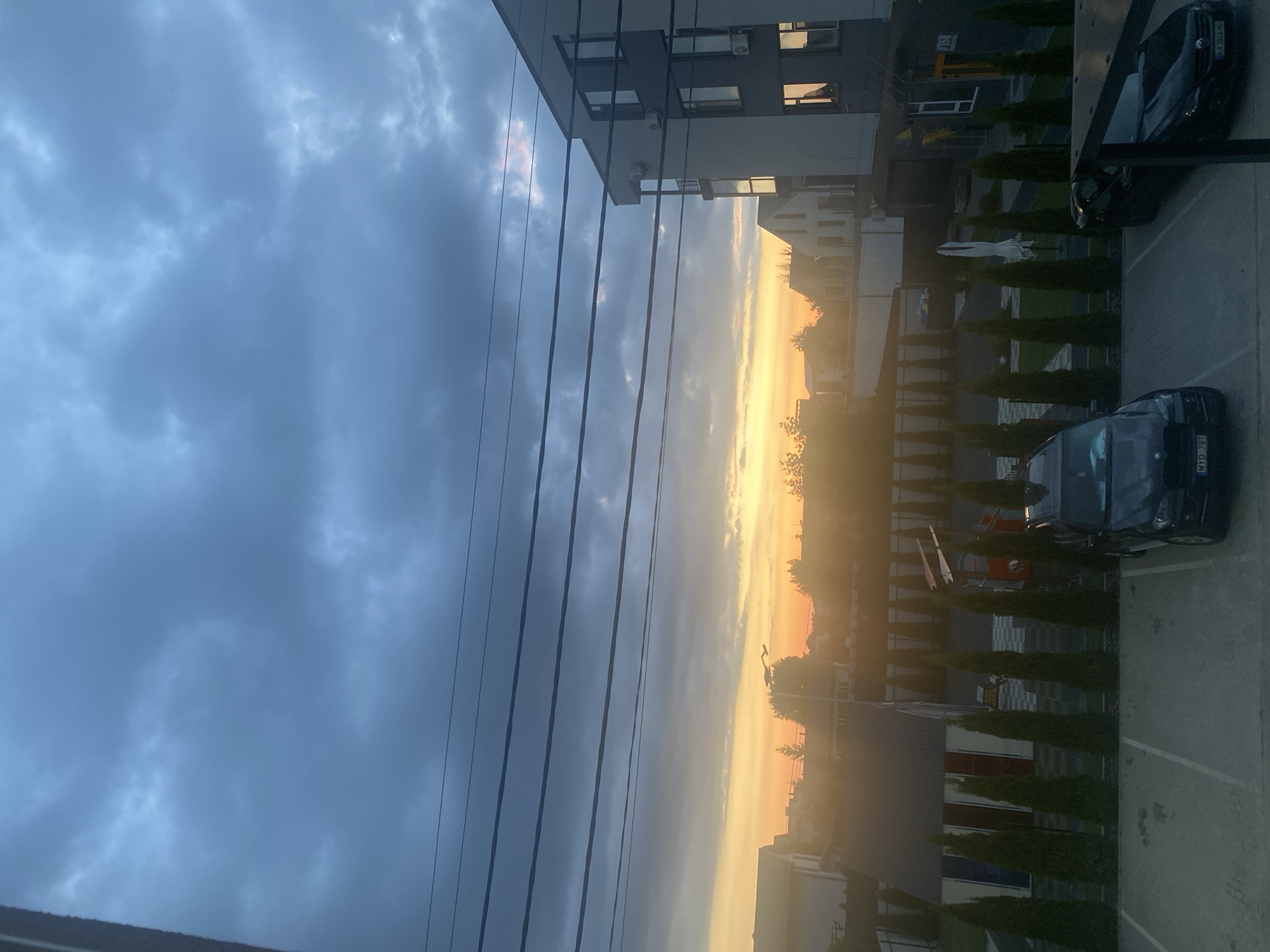 The image captures a view from a window looking out towards a residential area during what appears to be sunset or sunrise. The sky is filled with heavy, dark gray clouds, with a band of warm orange and yellow light peeking through the horizon, suggesting a transition period of the day. Several power lines are strung across the upper portion of the frame, obscuring some of the sky.
In the middle ground, there are several houses and buildings of varying architectural styles. One prominent building on the right is a modern, multi-story structure with a dark facade and white accents, featuring multiple windows and balconies. Adjacent to it, to the left, are smaller residential buildings with pitched roofs. A row of neatly trimmed evergreen trees forms a line in front of a low wall or fence. Further back, more houses and trees are visible, softened by the hazy light.
The foreground shows a parking lot with two dark-colored cars. The car on the left is a dark SUV with visible license plates. The car on the right is a darker sedan, also with a license plate. The parking lot is marked with white lines. The overall impression is a quiet, possibly suburban, scene as daylight fades or begins.
