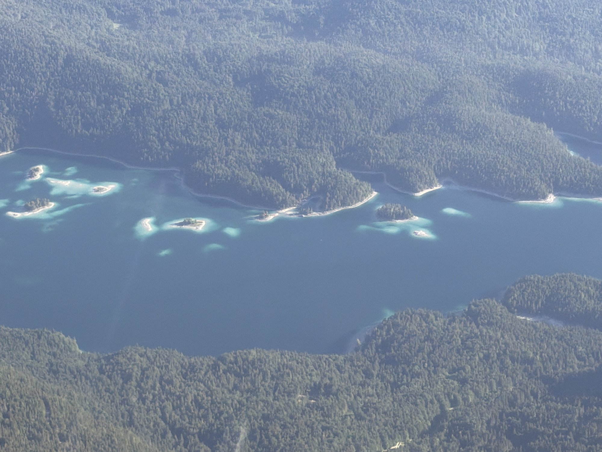 This aerial photograph shows the Eibsee lake in Grainau, Germany. The lake is surrounded by a dense, dark green forest, which covers the rolling hills. The water of the lake is a deep blue, with several small, tree-covered islands scattered throughout. The sunlight creates bright turquoise patches on the surface of the water, particularly around the islands. The overall impression is one of a serene and natural landscape.
