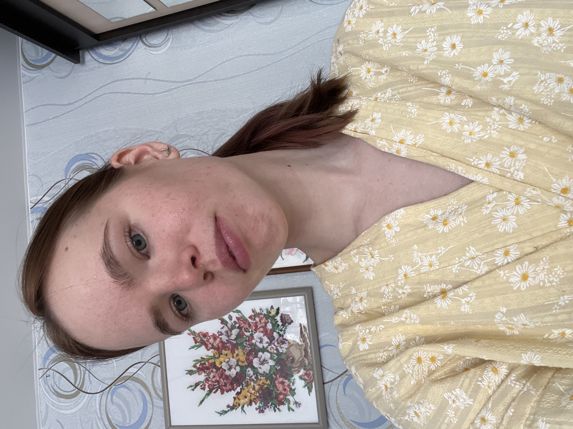 A young woman with auburn hair pulled back into a ponytail is shown in a close-up indoor photograph. She has fair skin, blue eyes, and full lips. She is wearing a light yellow dress with a white floral pattern. In the background, there is a wall with blue and white swirling wallpaper, and a framed floral print hanging on the wall behind her. The location is Chaikovsky, Russia.