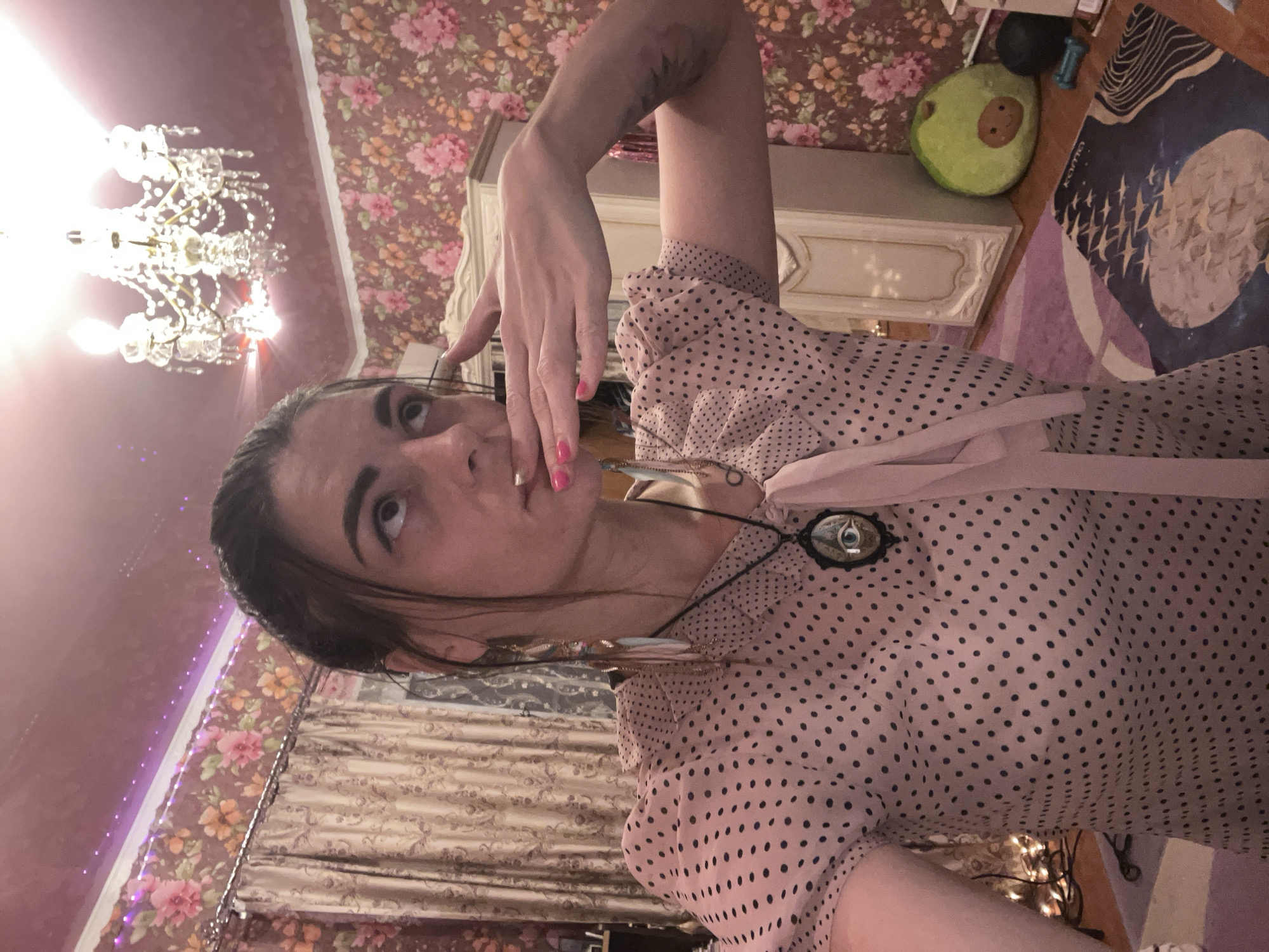 The image depicts an indoor scene, likely a bedroom, with a woman in the foreground. She has dark hair pulled back and is wearing a pink polka-dot blouse. Her right hand is raised to her mouth, and she is looking upwards. She is wearing a necklace with a pendant featuring an eye design. The background shows floral wallpaper, curtains, a chandelier, and a large cabinet. To the right, there is a stuffed avocado and some exercise equipment. The overall ambiance suggests a personal living space. The location provided, Krasnoe-na-Volge, Russia, is not visually discernible from the image itself.