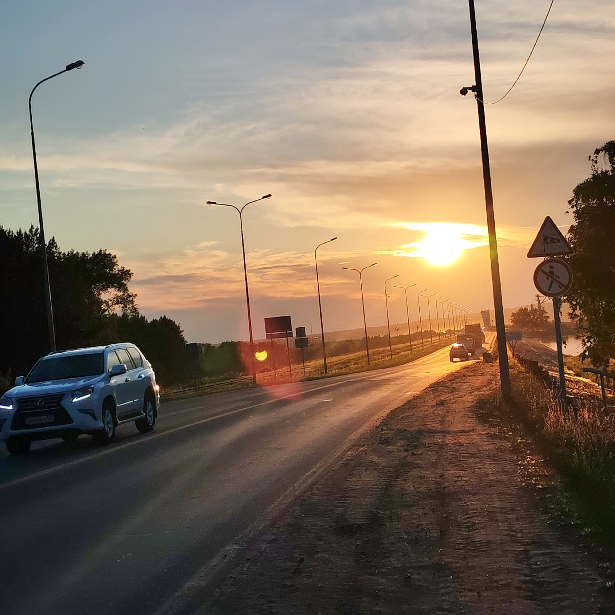 The image depicts a sunset scene on a road, likely a highway, with multiple streetlights lining the side. The sky is a gradient of blue and orange, indicating the sun is low on the horizon. A white SUV is driving on the road in the foreground. In the background, there are trees and distant structures. Traffic signs, including a warning sign for wind and a no-pedestrian sign, are visible near the road. The overall environment appears to be a rural or suburban setting on the outskirts of Chaikovsky, Russia, given the context provided. The road surface shows signs of wear and is slightly wet, reflecting the warm light of the setting sun.