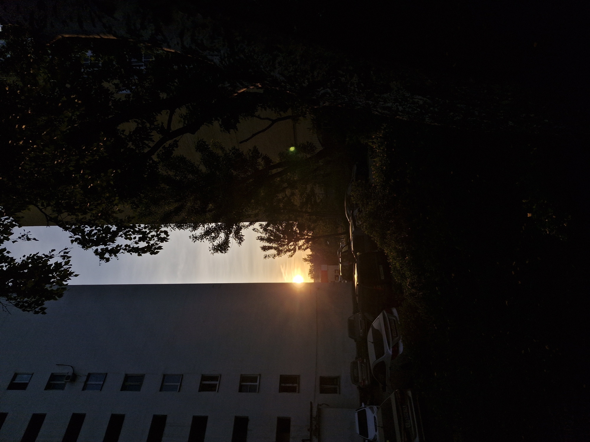 The image shows a view of a building at sunset in Yekaterinburg, Russia. The sun is positioned between the building and some trees, creating a silhouette effect. Cars are parked along the side of the building. The sky is visible above the building, transitioning from a lighter hue near the sun to a darker shade. The surrounding area is heavily shaded due to the sunset.