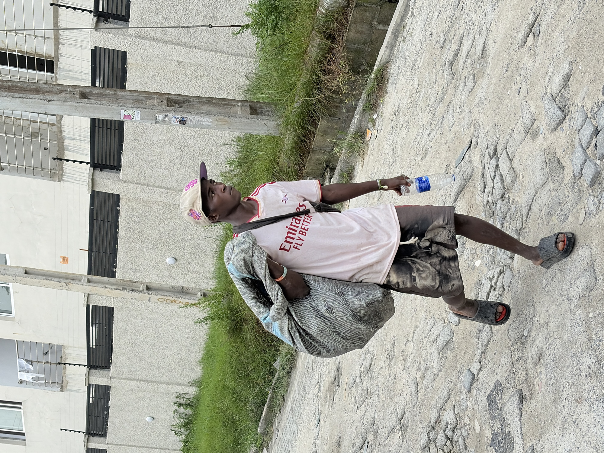 A young Black male wearing a white t-shirt with red trim and "Emirates Fly Better" printed on it, and patterned shorts, is walking on a dirt path in a semi-urban area. He wears a baseball cap and dark slides on his feet. He is carrying a large, rolled-up grey blanket over his left shoulder, and holding a plastic water bottle in his right hand. Lush green grass and a concrete drainage ditch are visible to his left, while a multi-story building with white walls and barred windows stands behind the grass. The ground is uneven and covered with loose stones and sand. The lighting suggests it is daytime, possibly late morning or early afternoon. The overall scene depicts a person going about their daily activities in what appears to be a warm climate.