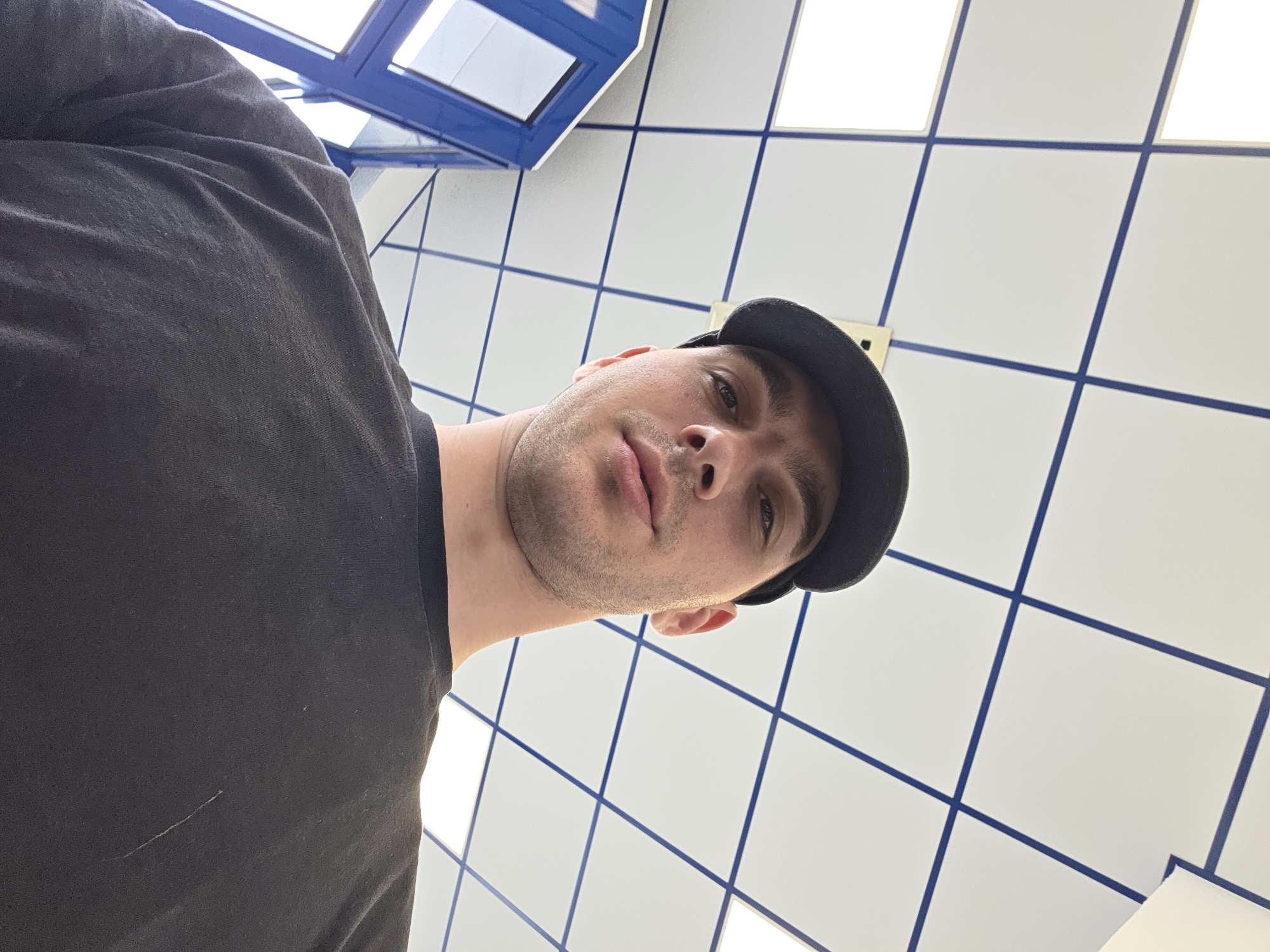A low-angle, medium shot shows a man wearing a black t-shirt and a black baseball cap, facing forward with his head tilted slightly back. He has short brown hair, brown eyes, and stubble on his chin. His mouth is slightly open. Behind him, a white tiled ceiling with blue grid lines is visible. To his left, a blue and white structure with windows is partially visible. The lighting is bright, suggesting an indoor environment.