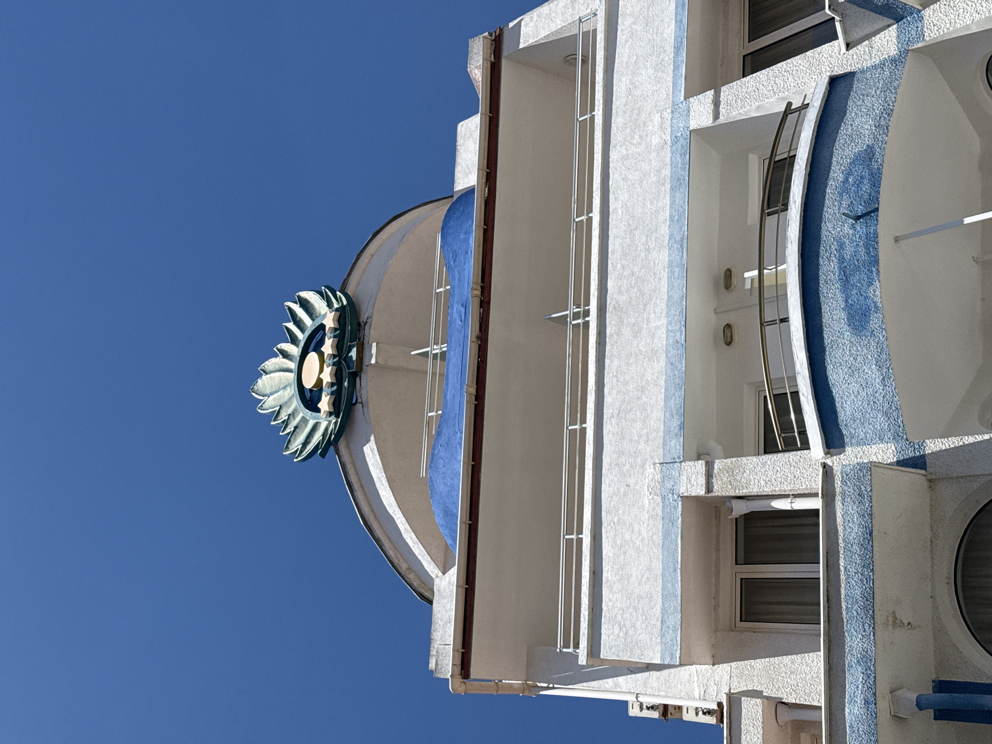 The image shows the upper facade of a building with white and blue detailing, located in the city of Sozopol, Bulgaria. The building has multiple balconies with metal railings and several windows. At the very top of the building, a decorative element resembling a stylized flower or sunburst is visible, with golden stars beneath it. The sky is clear and blue.