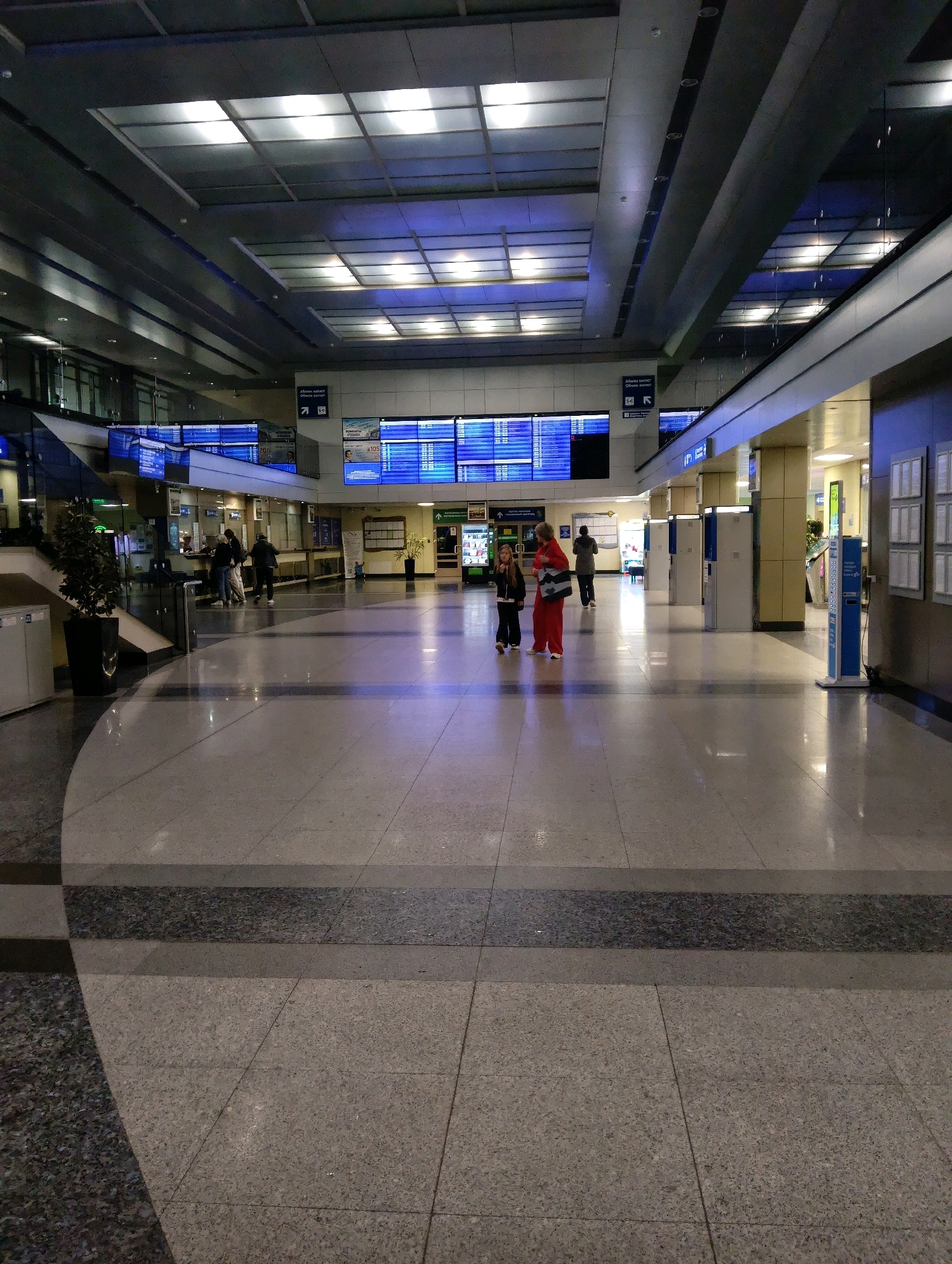 The image shows an interior view of a modern airport terminal. The ceiling features a grid of fluorescent lighting panels and a dark, angled structure. Large digital display boards with blue screens showing text and schedules are visible on the walls in the background. In the foreground, two people, a woman in a red outfit and a child in dark clothing, are walking. The floor is polished stone with a dark, speckled band running across it. Several kiosks, ticket counters, and information booths are present. The overall environment appears clean, well-lit, and functional. There is no visible information in the image that indicates this location is in Hrodna, Belarus.