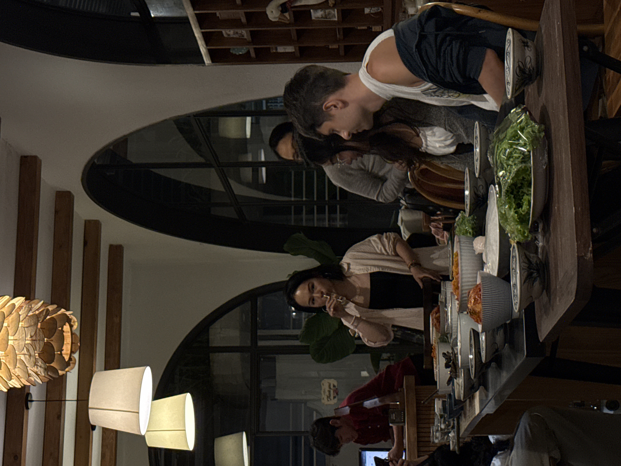 In the city of Lam Vien - Da Lat, Vietnam, a group of people are gathered around a dining table laden with food. The scene appears to be indoors, with warm, ambient lighting provided by decorative ceiling lamps. A woman in the foreground, wearing a light-colored open shirt over a black top, is drinking from a glass. To her left, a young man in a red shirt is looking at his phone. Behind them, other individuals are visible, including a person in a grey sweater and another man in a white tank top and a dark jacket. The table is filled with various bowls of food, including what appears to be kimchi and fresh lettuce, suggesting a communal meal. The background features arched doorways and wooden shelving units, adding to the cozy and inviting atmosphere of the dining space.