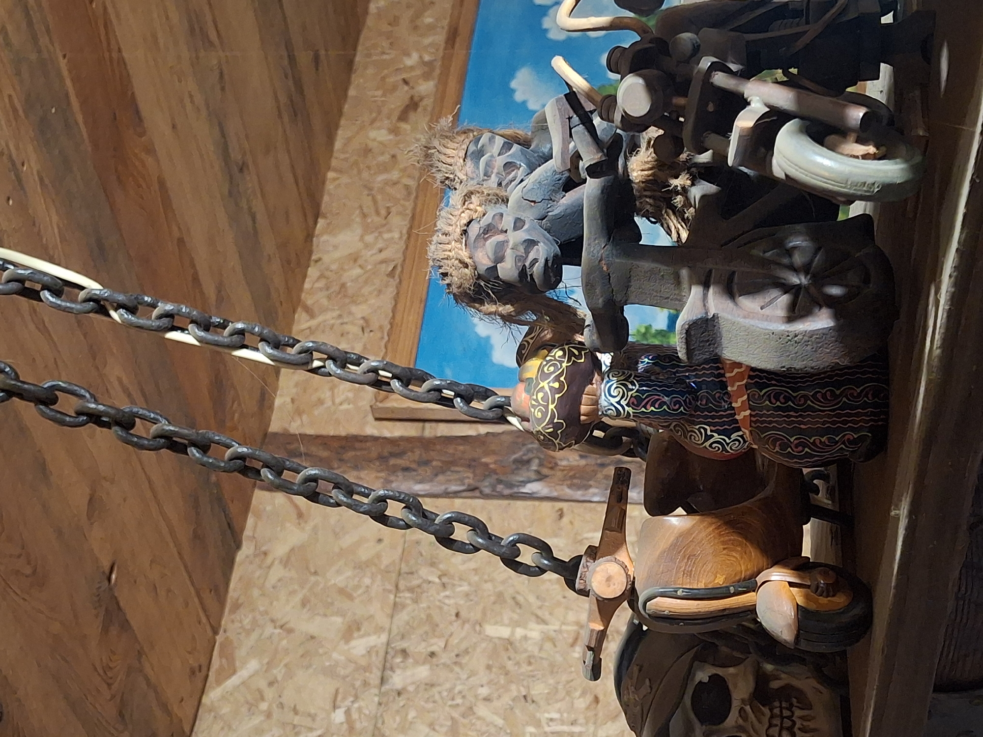 The image displays a collection of intricate wooden carvings and a heavy metal chain, set against a rustic wooden background and a painted sky mural. A dark, robust metal chain stretches horizontally across the left and center of the frame. Attached to the right side of this chain, a dense arrangement of carved wooden objects is showcased. These objects include elements resembling a stylized wooden motorcycle or vehicle, featuring detailed parts like wheels and structural components. Other carvings suggest parts of a saddle or harness, with some sections wrapped in natural, fibrous rope. Several pieces are adorned with elaborate patterns in red, black, white, and golden-yellow, showcasing detailed craftsmanship.
The setting appears to be an indoor display, possibly in a museum, a craft shop, or a themed interior. The background features a wooden wall, composed of light brown planks on the left and oriented strand board (OSB) on the right. Behind the wooden objects, a vibrant mural depicts a bright blue sky with white clouds, providing a contrasting, artificial outdoor element to the indoor scene.
No activity or event is taking place; the image captures a static, decorative arrangement. The time of day and weather are not discernible due to the indoor setting. The items appear to be well-lit, possibly by artificial light. While the location context is provided as Chaikovsky, Russia, there are no specific visual cues within the image to indicate this. There is no visible text in the image.