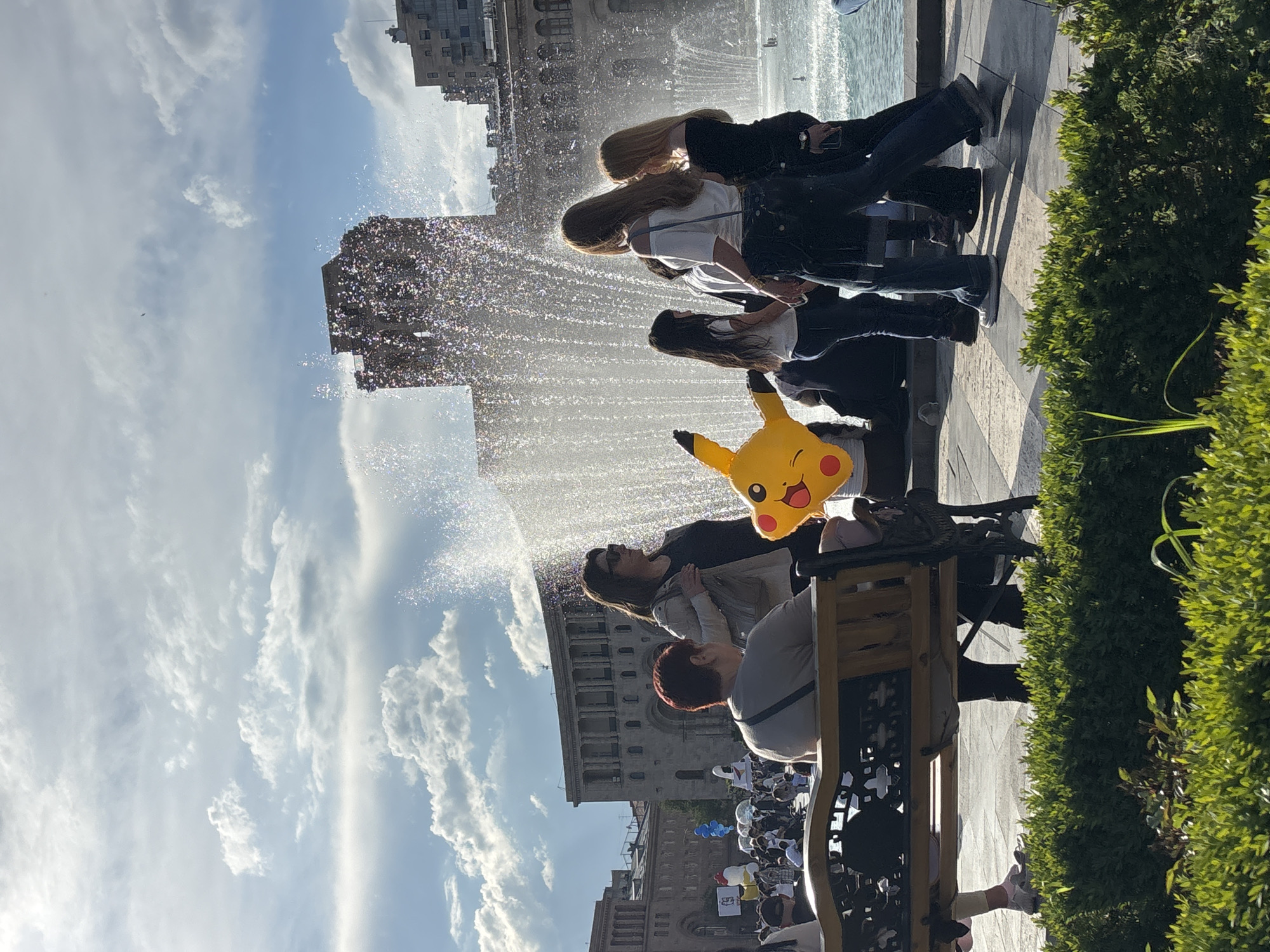 The image captures a bustling daytime scene in a public square in Yerevan, Armenia. A large fountain dominates the midground and left side of the frame, spraying a sparkling cascade of water illuminated by bright sunlight.
In the foreground and midground, several people are gathered, observing the scene. Most are women, seen from behind or in profile, with long hair. A large, cheerful yellow Pikachu-shaped balloon is prominently held by one person near the center-right. Another individual is seated on a decorative park bench, partially visible on the right. The ground is a patterned paved area, and lush green hedges line the right side of the composition.
In the background, under a bright blue sky with scattered white clouds, imposing buildings with classical architectural elements are visible. Further in the distance, a substantial crowd of people is gathered, indicating a larger public event or demonstration. Some members of the distant crowd are holding signs or banners, and colorful balloons (blue, white, red, yellow) can be seen among them, though the text on the banners is illegible.
The clear sky and strong illumination suggest it is a sunny day, likely late morning or afternoon. The overall atmosphere is lively, characteristic of a public gathering in a city square.