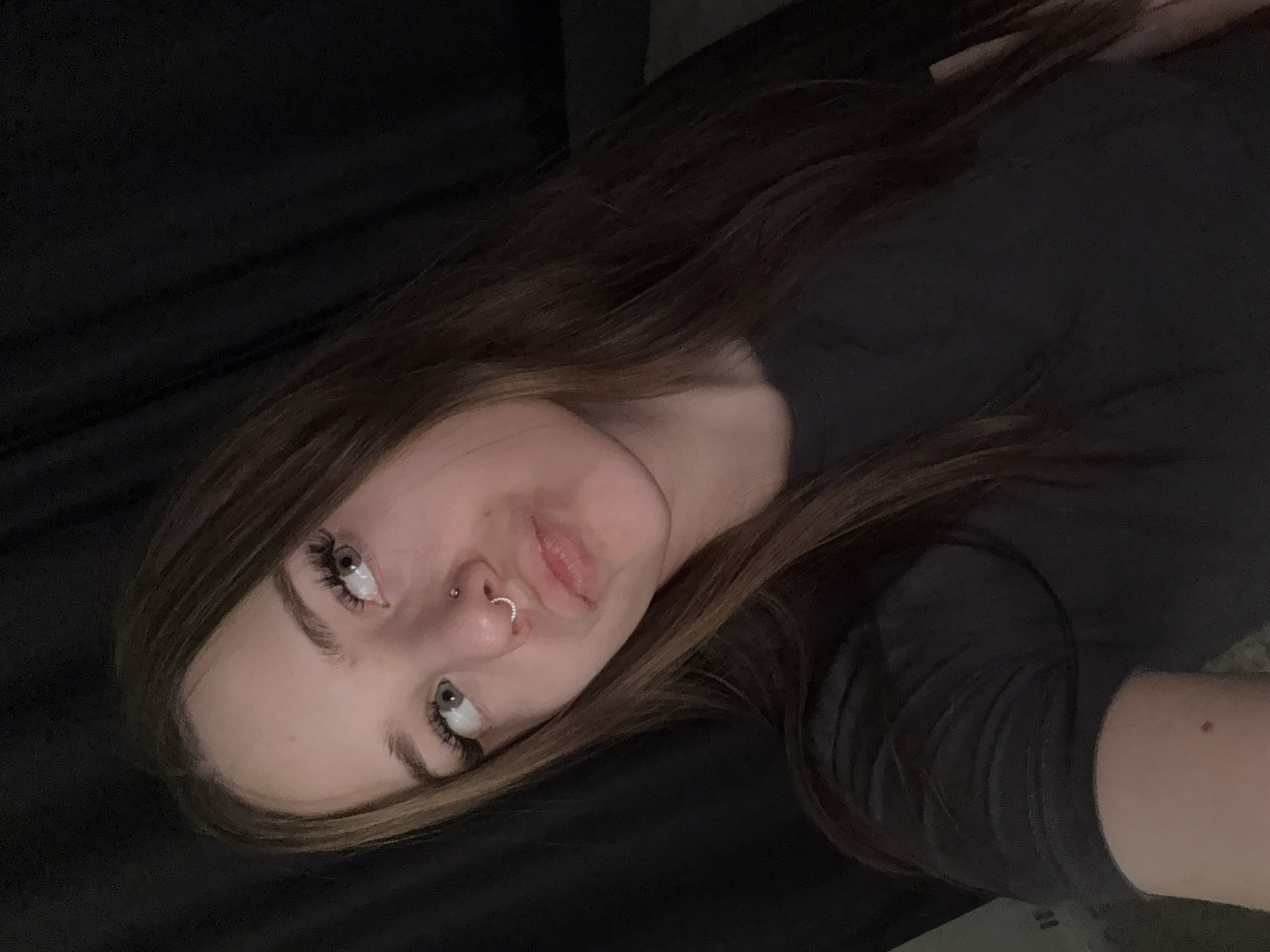 A young woman lies on a dark, soft surface, possibly a bed or couch, with her head tilted towards the camera. She has long, straight brown hair that falls across her face and shoulder. Her eyes are light-colored and look upwards. She has a nose piercing. She is wearing a dark-colored t-shirt. The background is out of focus and appears to be a dark, draped fabric. The photo has been taken at a close range. The location in the city of Chasovnya, Russia cannot be determined from the image.