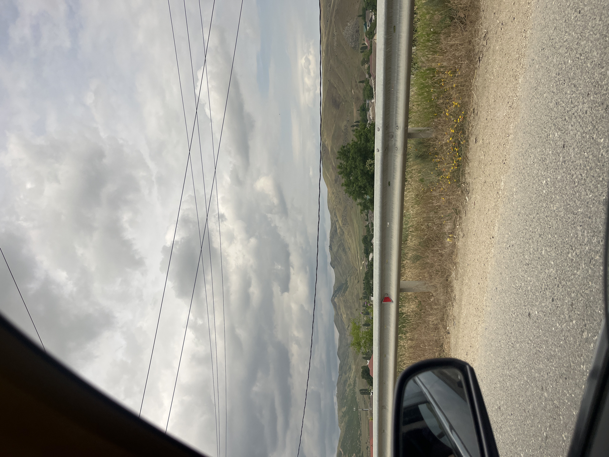 The image captures a view from inside a vehicle, looking out towards a landscape. The foreground shows a portion of the car's interior, including the side mirror and the asphalt road. A silver guardrail runs horizontally across the middle of the frame, separating the road from a dry, grassy embankment dotted with small yellow wildflowers.
Beyond the guardrail, a village or town is visible nestled at the foot of rolling, dry, grassy hills. The houses have reddish-brown roofs. The hills are undulating and appear arid, with sparse vegetation. In the background, the sky is overcast with dense, grey clouds. Several dark power lines are strung across the upper portion of the image, originating from the left side and extending towards the right. The overall scene suggests a rural or semi-rural setting under a cloudy sky, likely in the region of Karabudakhkent, Russia, given the context. There are no people or animals clearly depicted in the image.