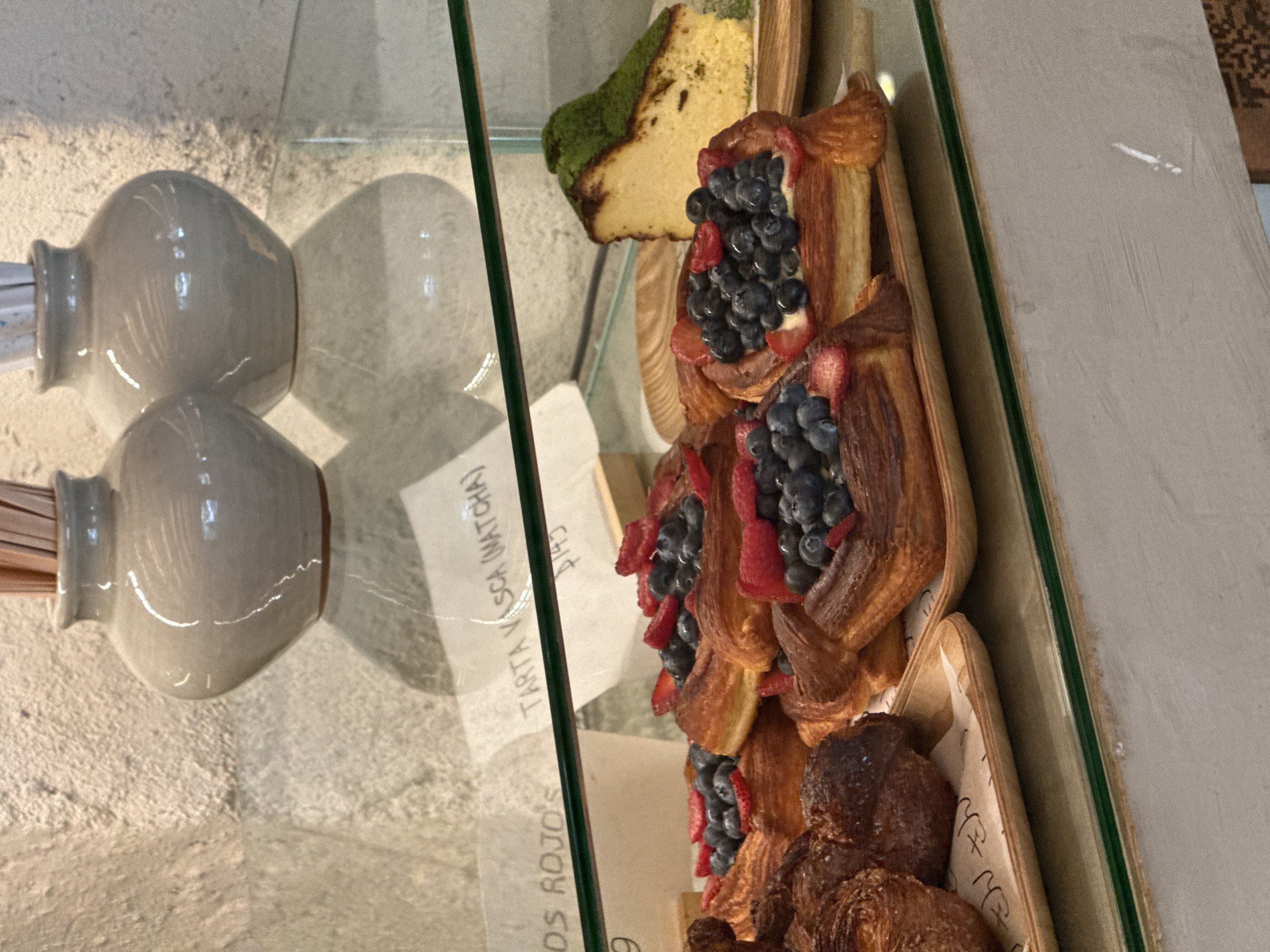 A bakery display case contains pastries and desserts, including a matcha cake slice and pastries topped with blueberries and strawberries, possibly in Mexico City, Mexico. Two ceramic containers with cylindrical objects inside are visible in the reflection. The pastries are priced and labeled, with "Tarta V. Scha (Matcha)" visible on a label.