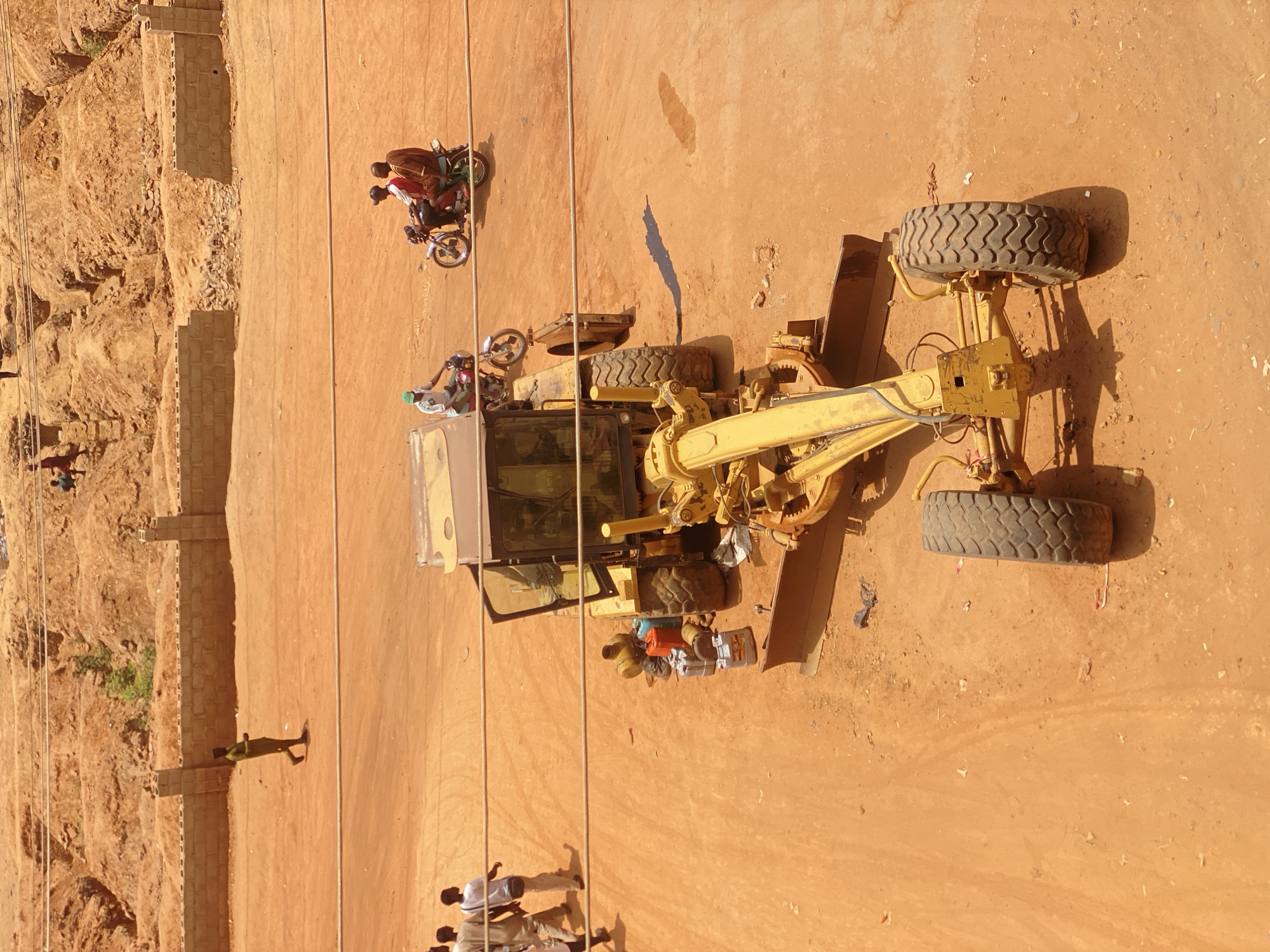 This is an aerial view of a section of Tudun Wada, Nigeria. A large yellow grader is positioned on a dusty orange-brown surface, likely a road or construction site. A motorcycle with two people is driving nearby. Several people are walking along the side of the road near a low brick wall. A second motorcycle with one person is parked on top of the grader. There are power lines extending from the top to the bottom of the image. The area appears arid with scattered vegetation.