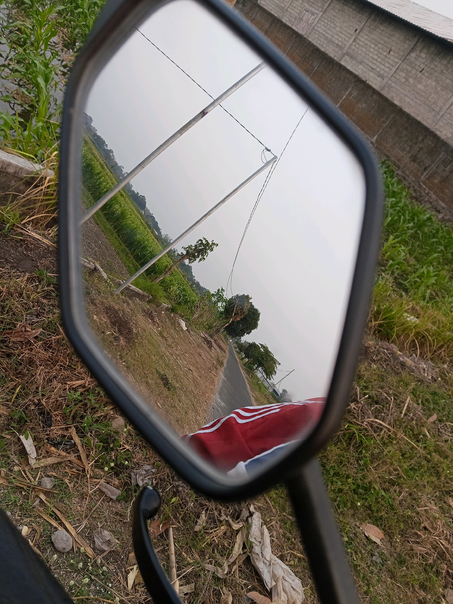 The image is a close-up, angled shot of a motorcycle's rearview mirror, reflecting a scene in Gogodeso, Indonesia. The mirror displays a landscape featuring a rural road passing through agricultural fields with green crops, likely rice paddies or corn. Several utility poles with wires are visible in the reflection. The background shows a long, brick building with a corrugated metal roof, possibly a warehouse or factory. In the foreground of the actual image, outside the mirror's reflection, is the ground covered with dry grass and some debris, along with a portion of a red and white striped object, which appears to be fabric. The overall lighting suggests it is daytime, likely late afternoon given the muted sky.