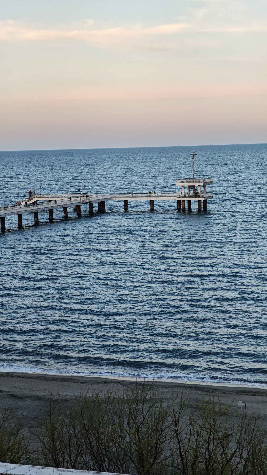 The image depicts a pier extending into the sea in Burgas, Bulgaria. The sea has slight waves. The sky above is pale pink. The pier is light-colored with dark support pillars and several people walking along its length. At the end of the pier is a platform structure with a tall post. Below the sea and pier, there is a sandy beach, followed by some sparse vegetation in the foreground. There's also a white chair on the beach.