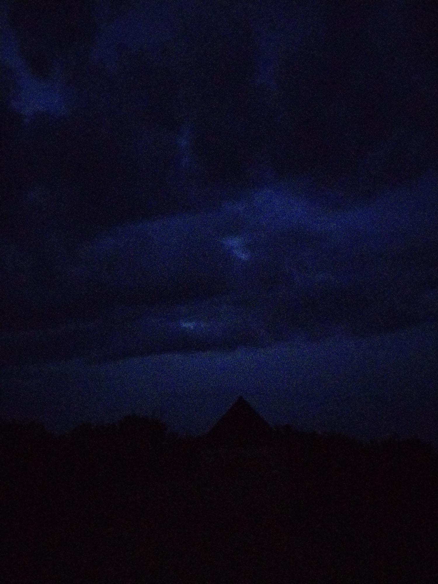 A silhouette of a pyramidal structure is visible against a dark, cloudy night sky. The structure appears to be a building or monument, possibly a church or a mausoleum, given its shape. The foreground consists of a dark, indistinct landscape, likely trees or rolling hills, creating a stark contrast with the lighter sky. The sky itself is a deep, dark blue, with lighter patches suggesting the presence of clouds illuminated by an unseen light source, possibly the moon or distant city lights. The image has a grainy texture, suggesting it was taken in low light conditions or with a camera that has a high ISO setting. No people or animals are visible in the scene. The location is identified as the city of Ustyvytsia, Ukraine, however, this information does not manifest visually within the image itself.