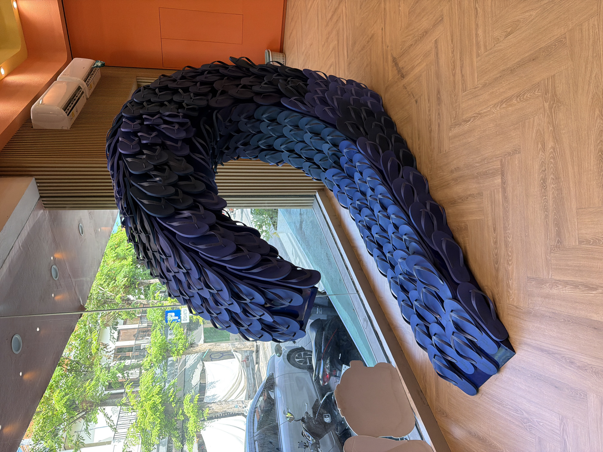 An indoor display in Kuta, Indonesia, consists of an arch-shaped structure made entirely of blue flip-flops. The floor is covered in brown wood-patterned tiles. The display is positioned near a glass window, and reflected in the glass window is the outside environment. Outside are trees, buildings, a white minivan, and a scooter. A blue parking sign is also visible. Inside the building, near the ceiling, are two air conditioning units. The ceiling is orange.