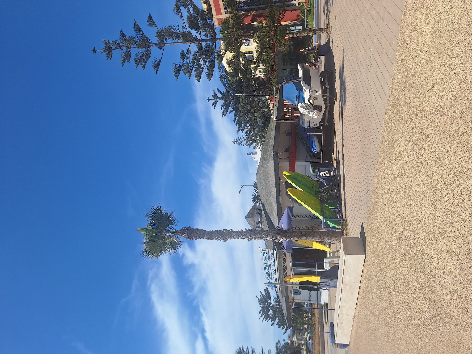 The image shows a bright, sunny day in Essaouira, Morocco, near the beach. In the foreground, there is a textured sandy area, transitioning to a paved walkway with geometric patterns. A long, light-colored concrete bench sits on the edge of the walkway. Behind the bench, a low-rise building with a covered outdoor area is visible. Several surfboards, two yellow and two green, are propped up against the building. A palm tree stands tall next to the building. Further back, a white boat on a trailer is parked. Beyond the building, several tall evergreen trees, characteristic of the region, are present, along with a multi-story building with red and yellow accents. The sky is a clear blue with scattered white clouds. Two individuals are walking in the background, near the multi-story building.
