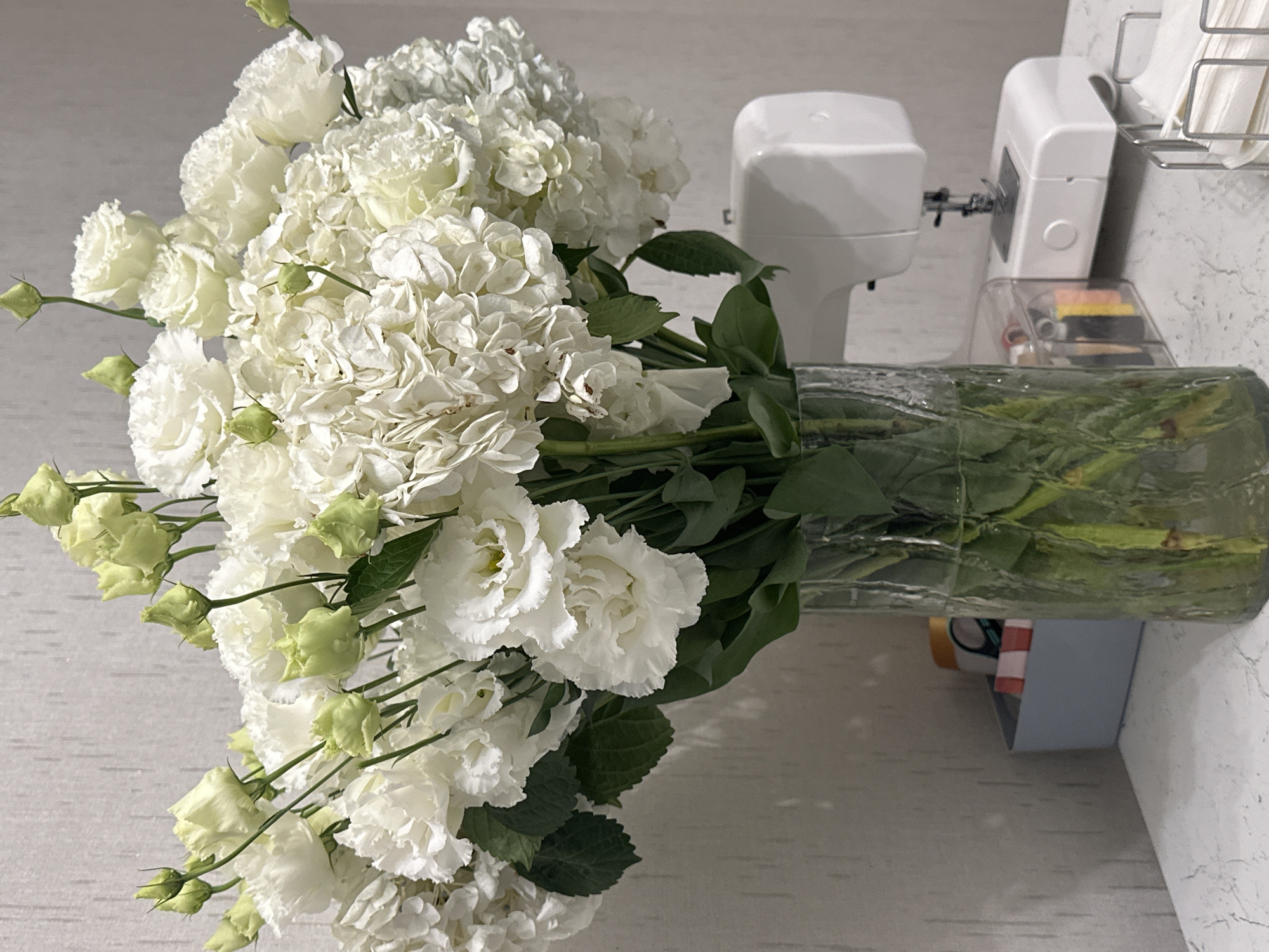 The image depicts a close-up, eye-level, indoor still life. The primary subject is a large bouquet of white flowers, predominantly lisianthus and hydrangeas, arranged in a clear, textured glass vase. Several buds are visible among the blooms. To the right of the vase, a white sewing machine is positioned on a white countertop with a marble-like pattern. In the background, a grey, textured wall serves as the backdrop. To the left of the vase, a small, light blue storage bin contains assorted colorful items. The scene is evenly lit, with soft shadows suggesting ambient light. There are no people or animals present. The image does not contain any information that would allow for determination of its location in Krasnodar, Russia.