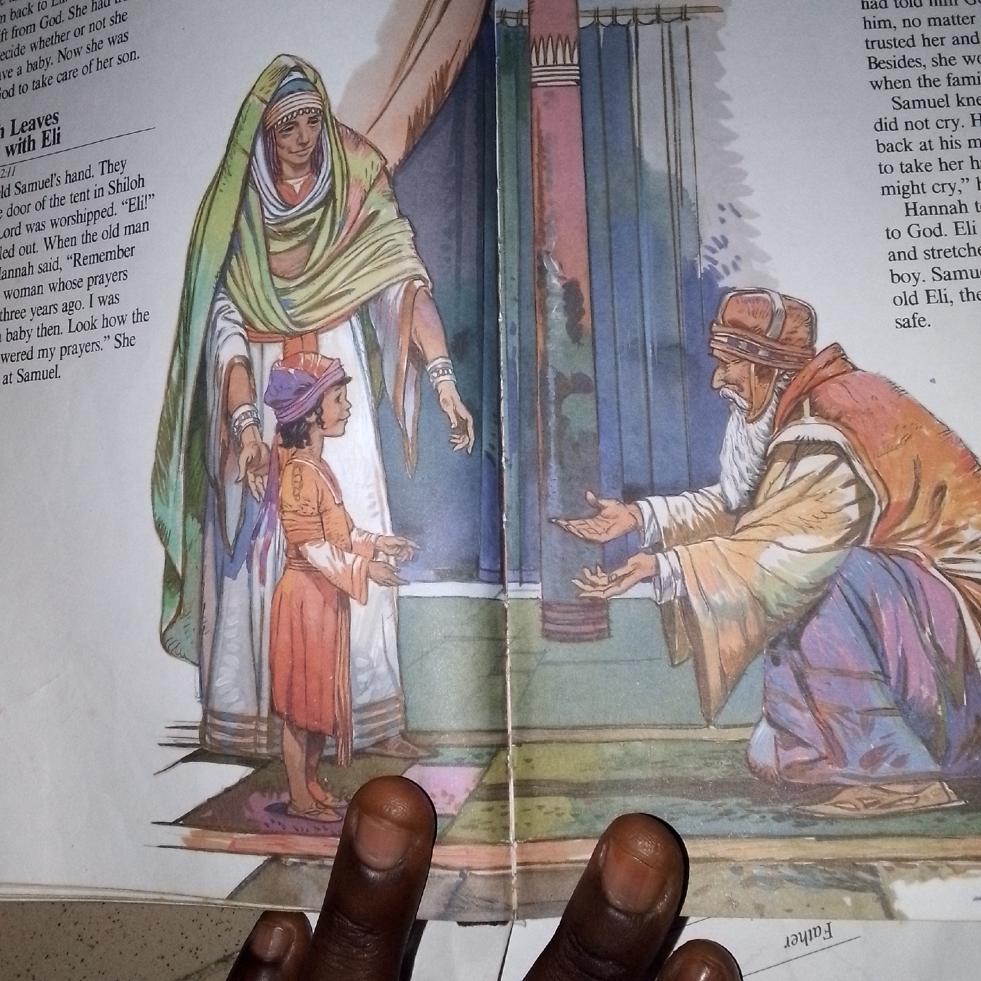 This is a close-up, eye-level, overhead shot of an open book, featuring an illustration depicting a biblical scene. In the illustration, a young boy, Samuel, stands between two adults. To the left, an older woman, possibly Hannah, stands behind Samuel, wearing a green head covering and a white robe. To the right, an older man, Eli, with a white beard and wearing a brown and orange robe, kneels, reaching out his hands towards Samuel. Samuel is dressed in an orange tunic and a purple head covering. The background of the illustration shows architectural elements resembling columns and curtains, suggesting an interior setting like a temple. The book itself is open, with text visible on both pages. On the left page, the text is a narrative about Samuel. On the right page, text is also visible, partially obscured by Eli's figure. The foreground of the photograph shows two dark-skinned thumbs and fingers from the photographer, obscuring parts of the bottom of the book and illustration. The lighting appears to be natural and even. The image does not contain any recognizable landmarks or features that would indicate its location in Jalingo, Nigeria.