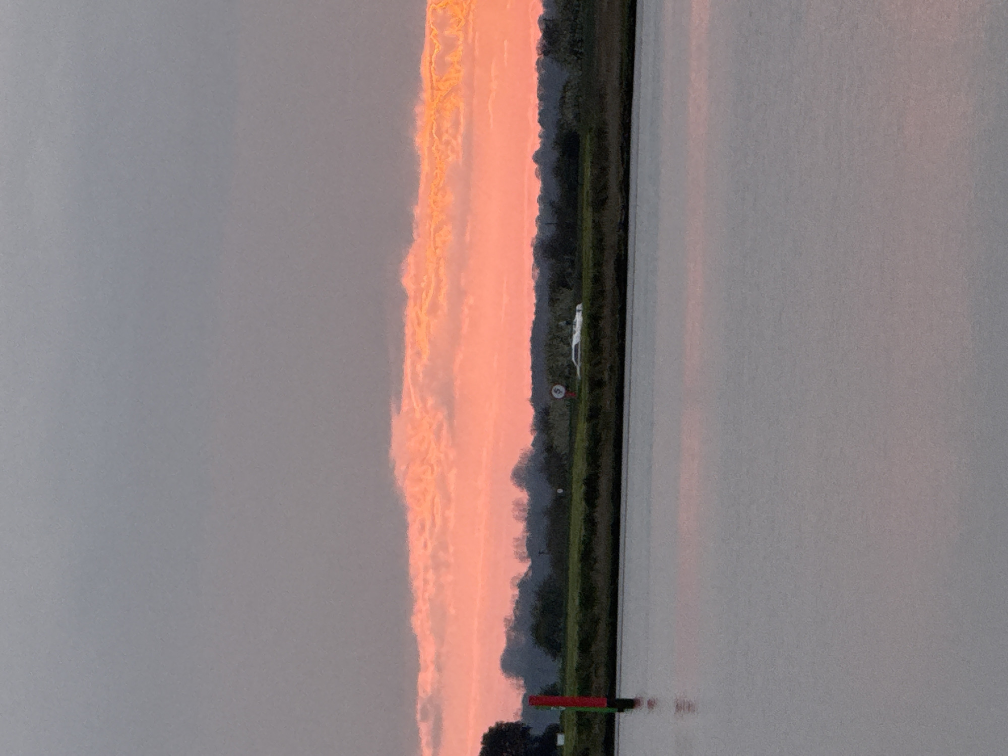 The image shows a body of water reflecting a pink and orange sunset on the right side of the frame. On the left, a gray sky is visible. In the center, a strip of land with green vegetation and trees divides the sky and water. There is a white object visible on the land strip, along with other small objects. In the bottom center, a red and green post stands in the water. Without further detail, it is difficult to confirm this is Great Yarmouth, United Kingdom, as it needs some reference to a local landmark.