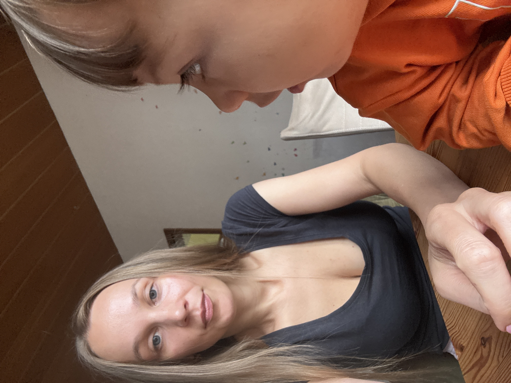 In Überherrn, Germany, an adult woman with long blonde hair and a black t-shirt is seated at a wooden table, facing forward. To her right, a young child wearing an orange shirt is partially visible, facing the woman. The room features a wooden ceiling, light-colored walls with colorful speckled decorations, and a white chair in the background. The woman's expression is neutral, and the child's mouth is slightly open, suggesting interaction or speech. The lighting is natural, casting soft shadows.